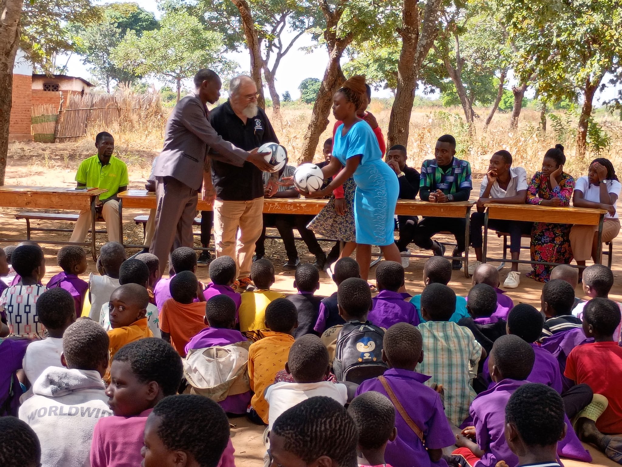 A woman in a blue dress receives soccer balls from two men during an outdoor event under trees, while children sit and watch.