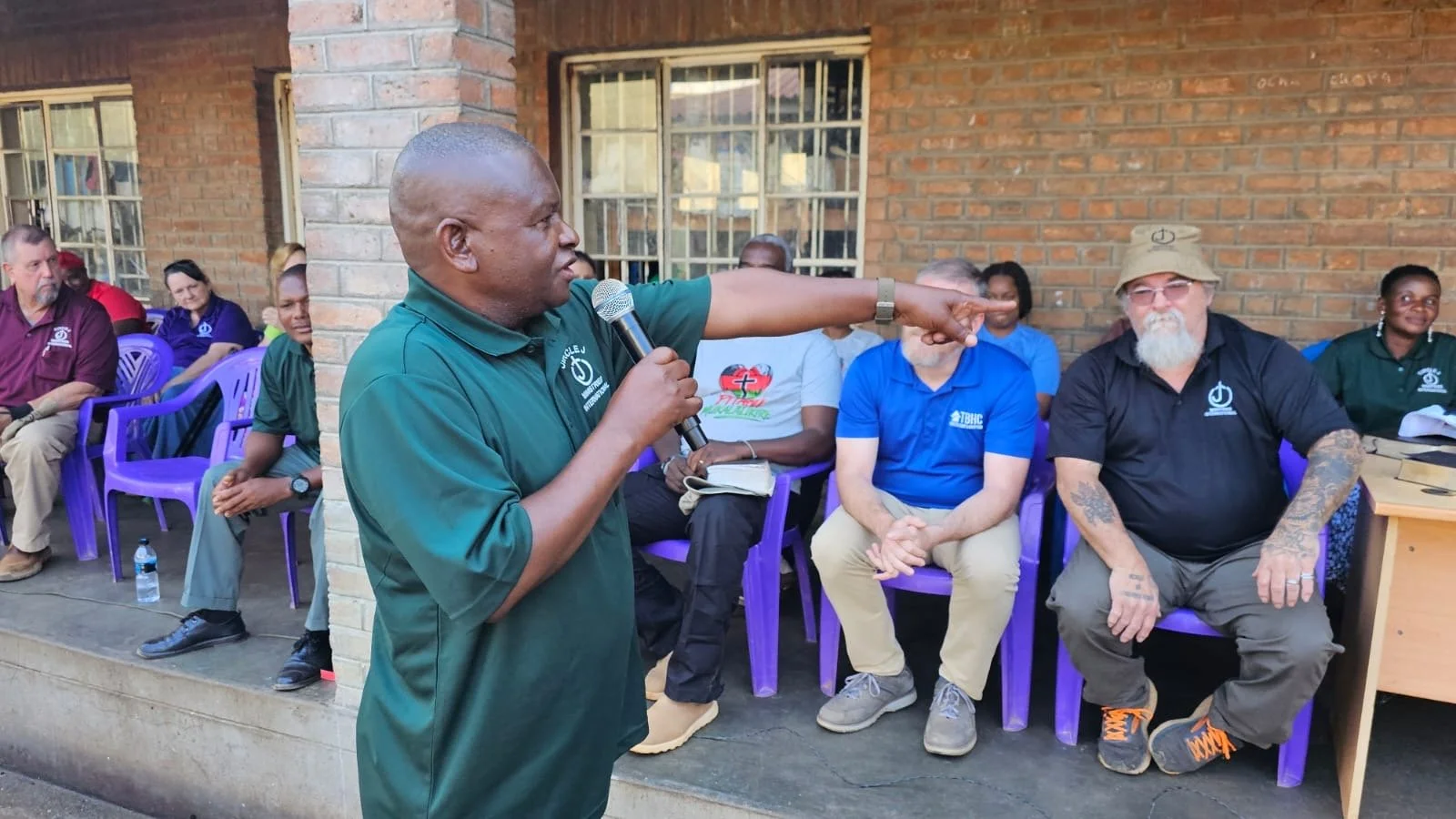 A man in a green polo shirt is speaking into a microphone and pointing with his right hand during an outdoor meeting. Several seated people, including a man with a beard and tattoos, are listening. The background features a brick wall with windows and purple chairs.