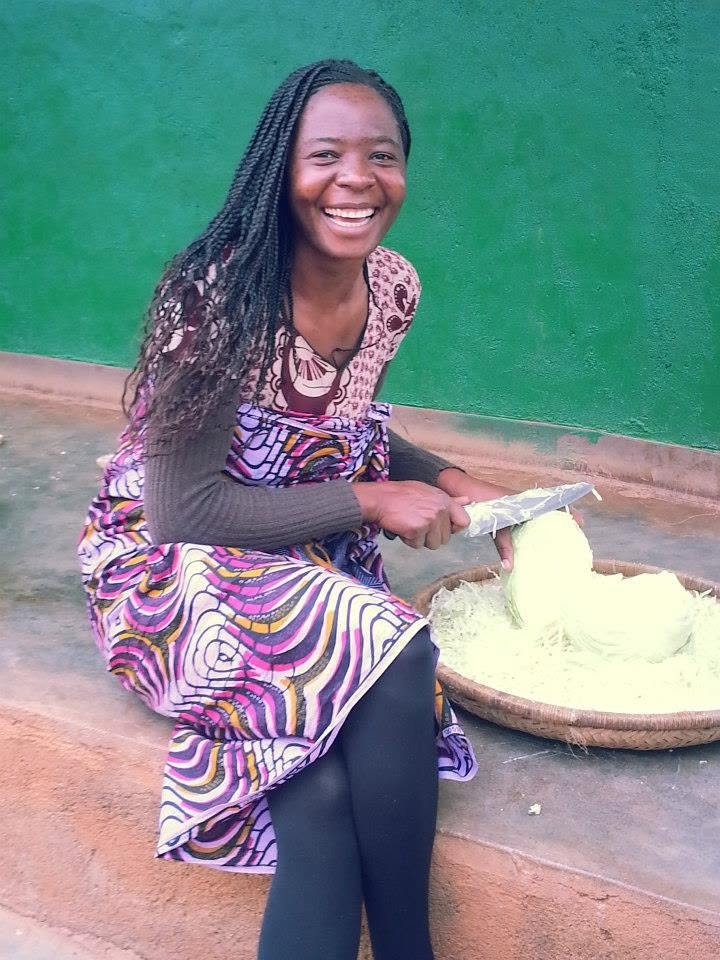 A woman with long braids smiling while sitting on a ledge, peeling raw cassava root into a large woven tray.