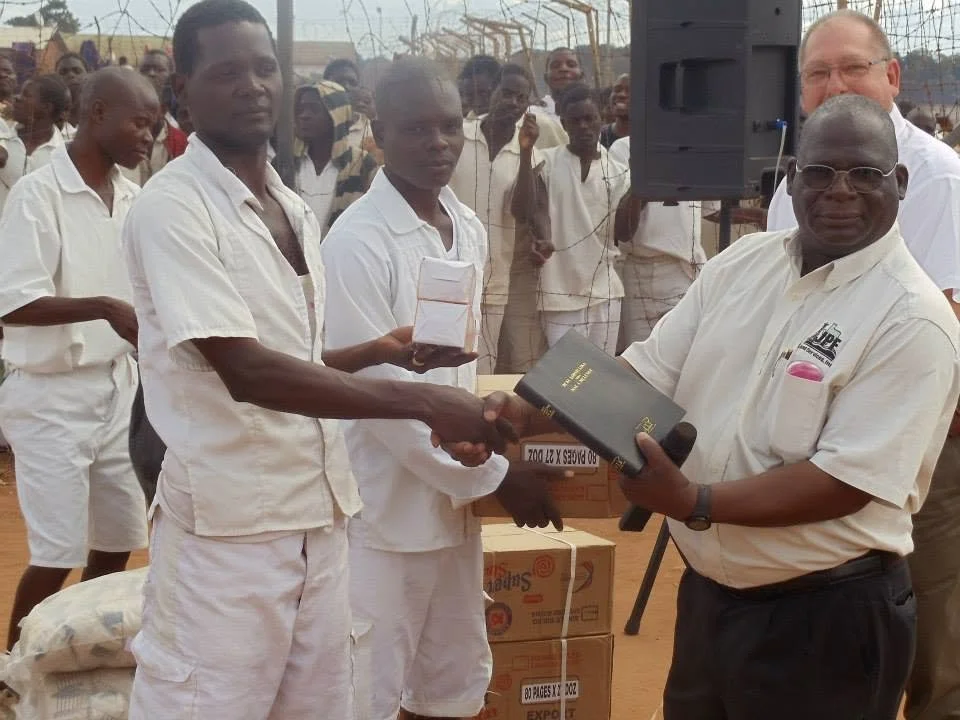 A group of men in white clothing at an outdoor event. One man is shaking hands with another, who is holding a black box. Several people are watching in the background, with boxes and bags on the ground nearby.