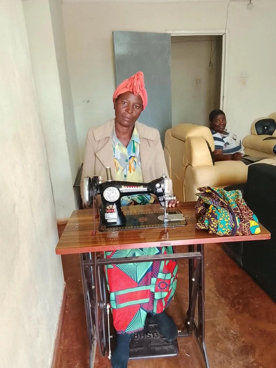 A woman sitting at a wooden table with a sewing machine, wearing a pink head wrap, beige jacket, colorful dress, and black shoes. In the background, another woman is seated on a beige sofa, smiling, wearing a striped shirt. The room has plain white walls and a brown floor.
