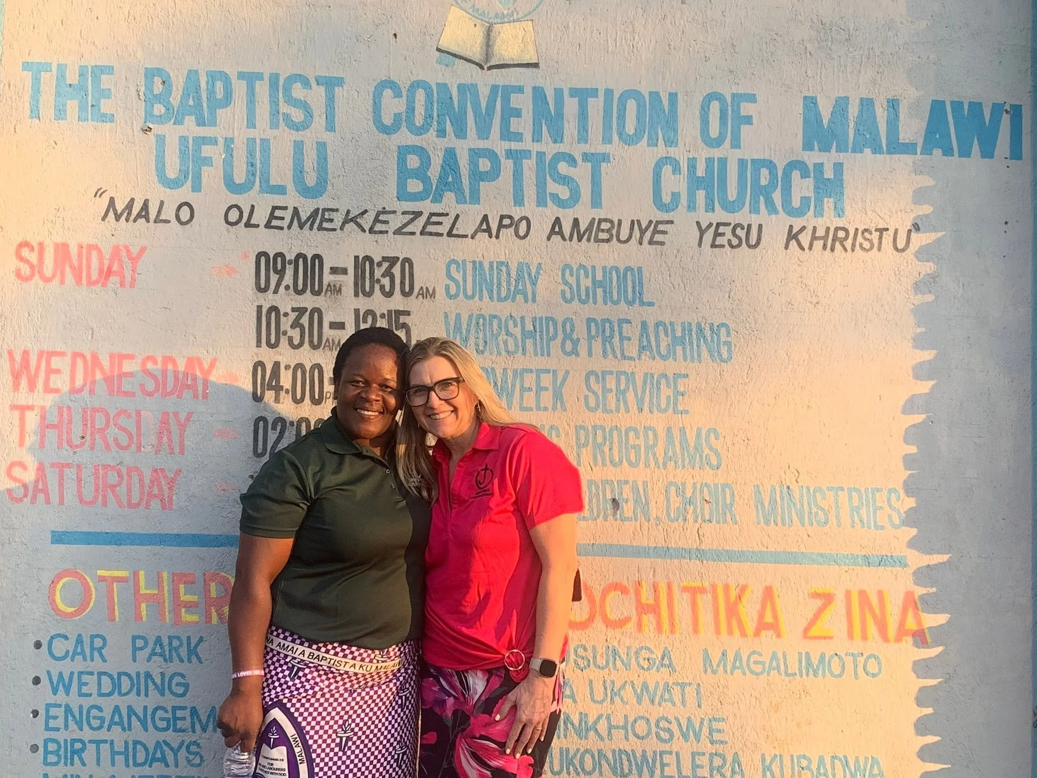 Two women smiling and standing in front of a large schedule board with blue and red text.