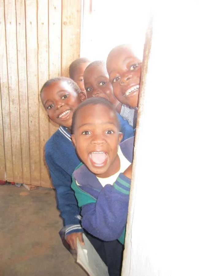 Six children peeking through a gap in a wooden wall, smiling and looking excited.