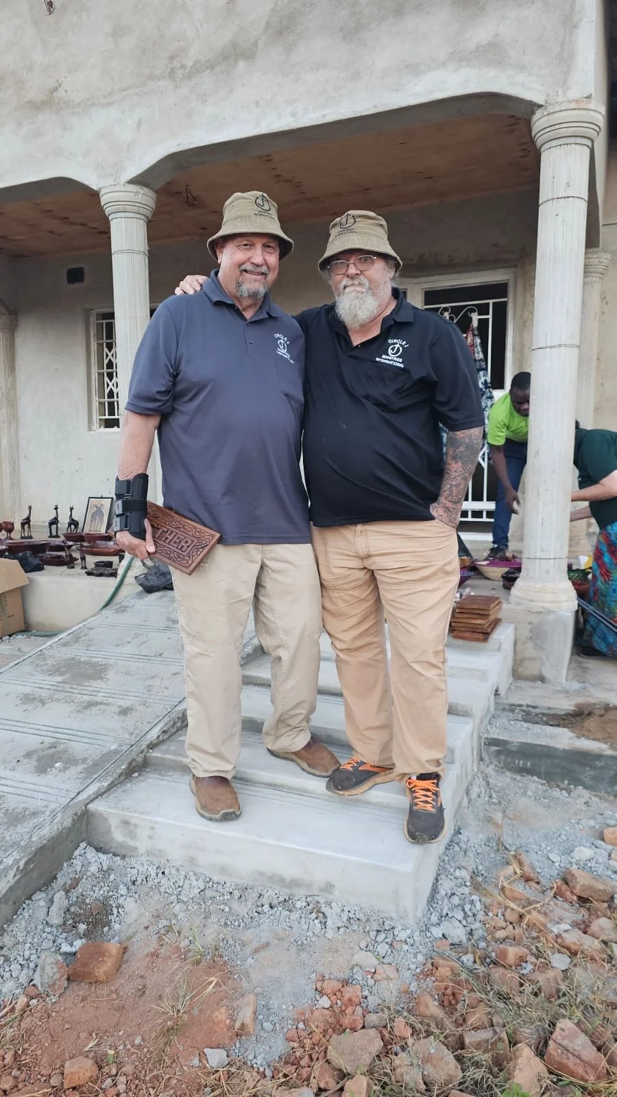 Two men standing on a porch of a house under construction. They are wearing hats and casual clothes, with other construction workers visible in the background.