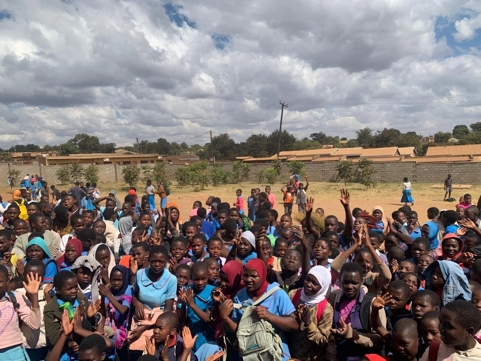 A large group of school children gathered outdoors, some raising their hands, with a wall, some trees, and buildings in the background under a cloudy sky.