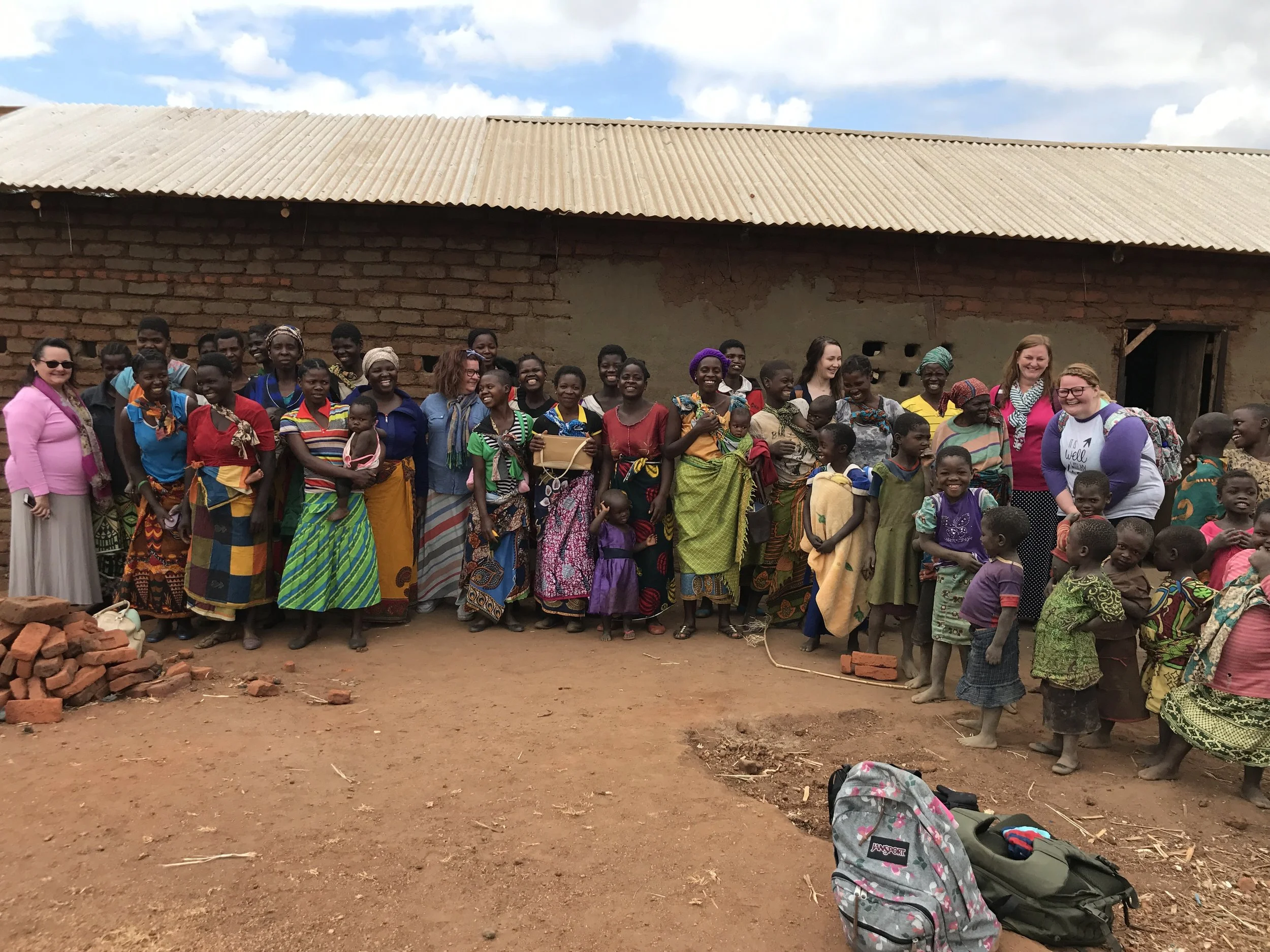 A large group of children and women standing outside a brick building with a corrugated metal roof. They are smiling, and some are holding children. There are backpacks on the ground in front of the group, and the ground is dirt.