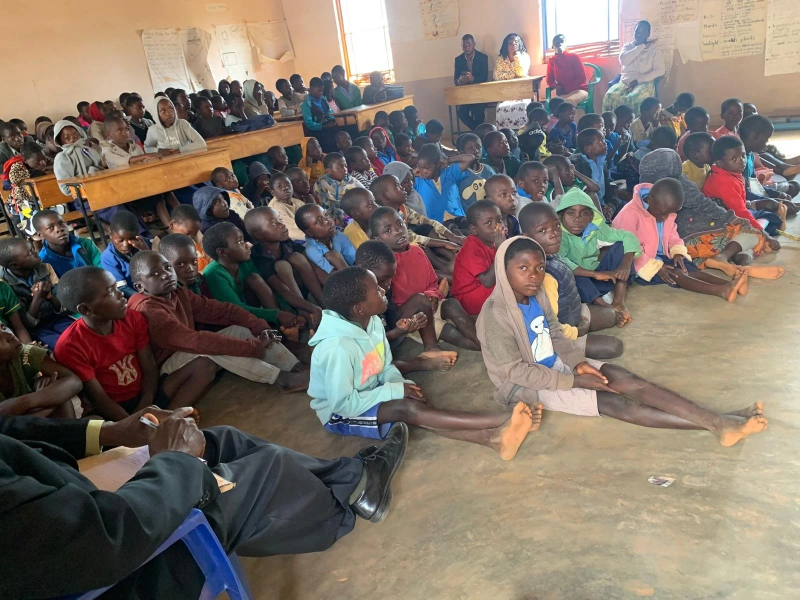 A large group of children sitting on the floor and on benches inside a classroom, attentively listening. Some children are seated with legs stretched out, others with knees up. There are a few adults at the back of the room near the windows.