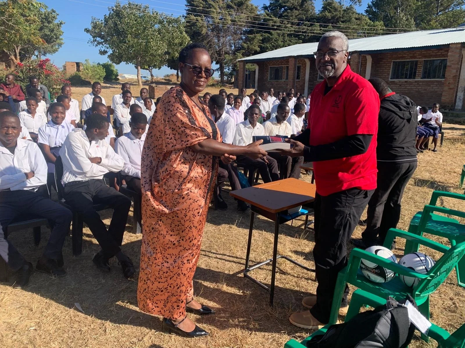 A woman in an orange patterned dress is receiving a book or award from a man in a red shirt during an outdoor ceremony, with students seated in the background.