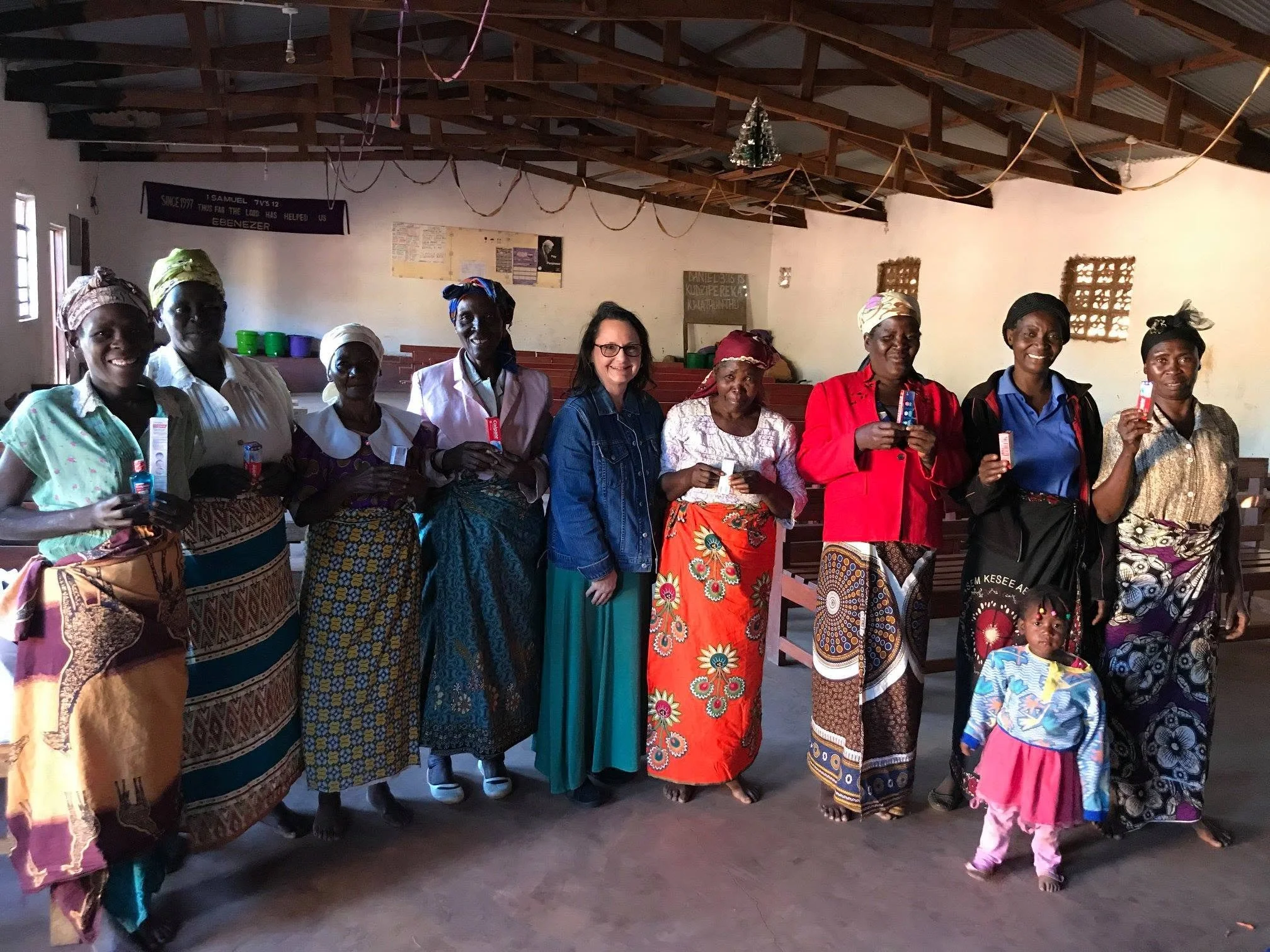 A group of women and a young girl standing inside a church or community hall, with some dressed in traditional colorful attire and headscarves, holding small items, against a backdrop of plain walls and wooden ceiling beams decorated with streamers and small Christmas tree ornament.