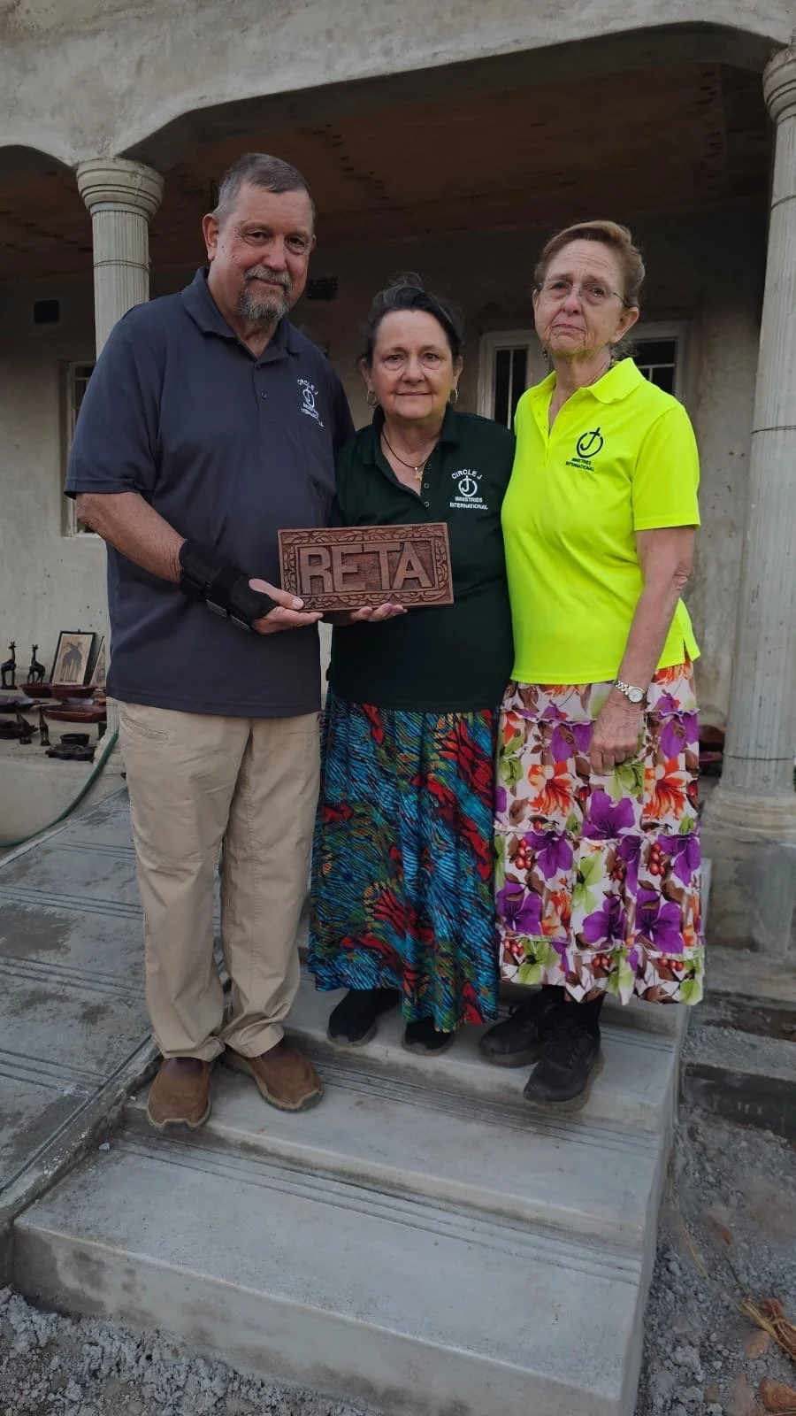 Three people standing on a stone step outside a building with columns, holding a wooden sign that reads 'RETA', with decorative carvings.