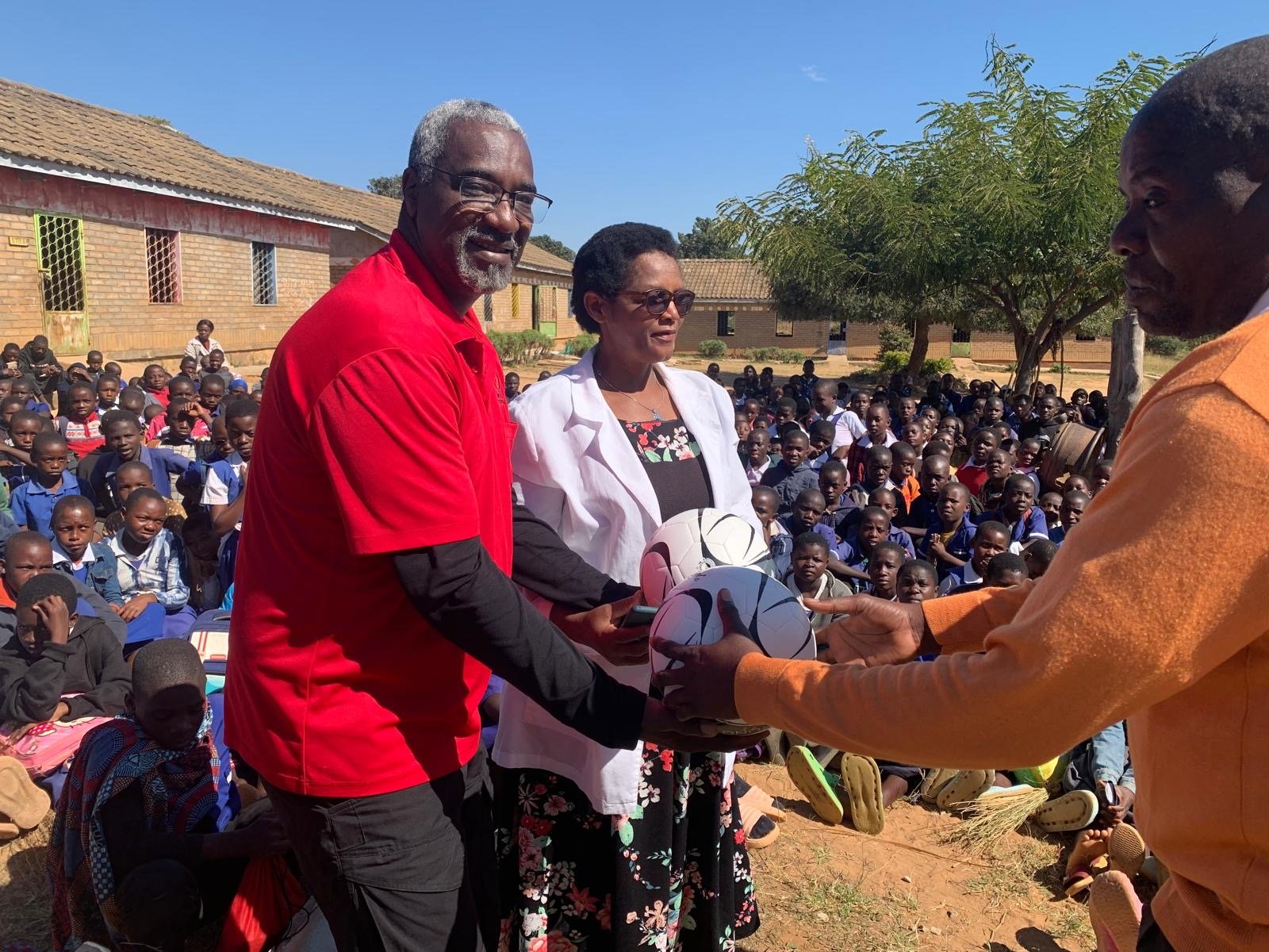 A man and a woman are standing outdoors, holding a football and receiving a token from another man during a school event. Many students are sitting in rows behind them, watching the scene take place.