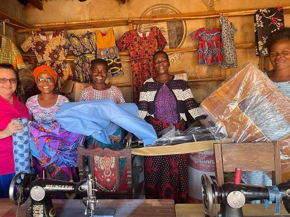 Group of five women in a sewing or craft shop, surrounded by colorful fabrics and garments, with sewing machines on a wooden table.