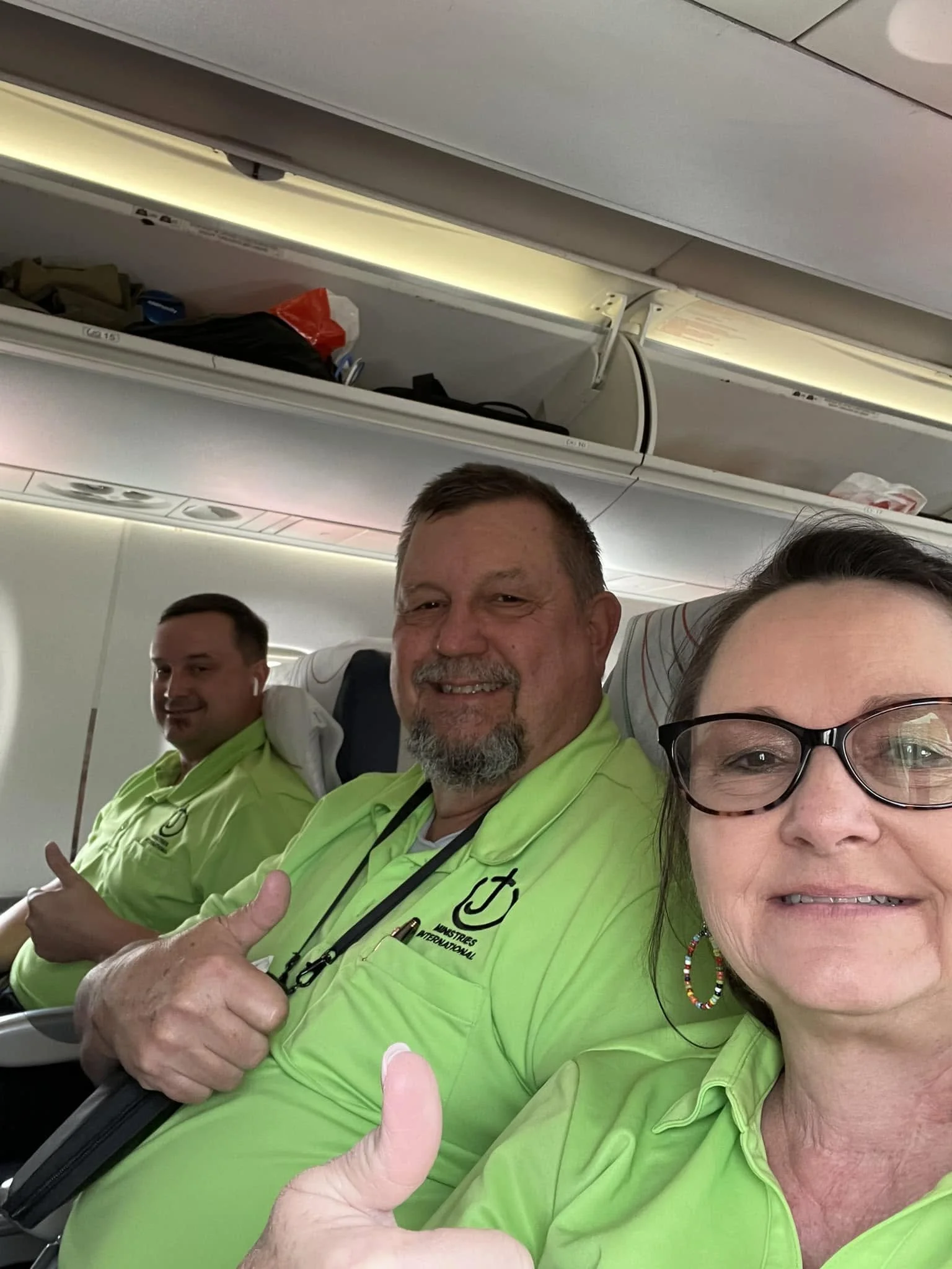 Group of airline crew members on an airplane, wearing neon green uniforms, smiling and giving thumbs up to the camera.