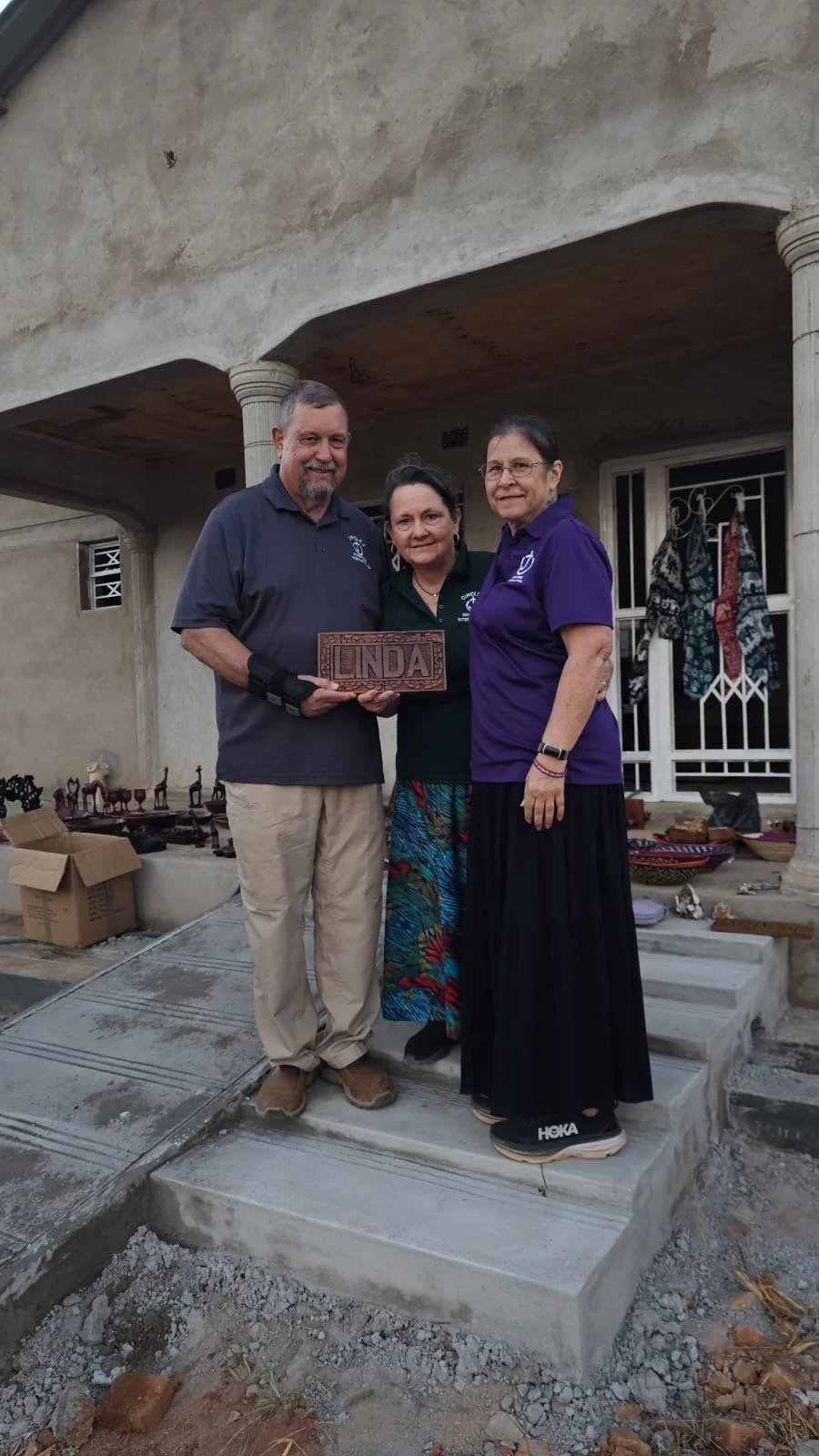 Three people standing on a concrete porch holding a carved wooden sign that says 'Linda'. Two women and one man, with the man wearing a black wrist brace. They are outside a house with various decorative items and boxes visible nearby.