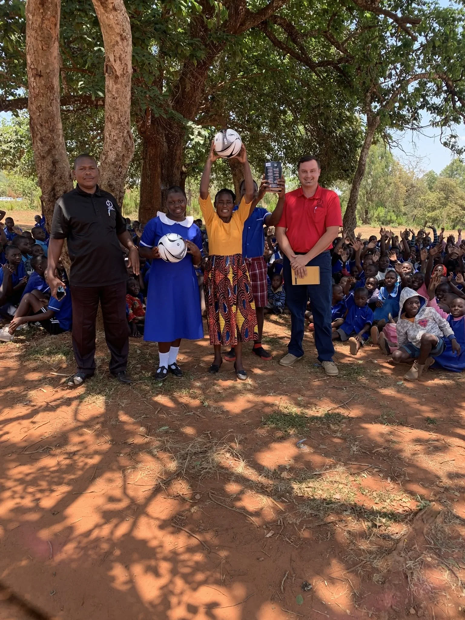 Group of children and two adults outdoors under a large tree, with one girl holding a soccer ball and another holding a book, while others sit and stand around in a sunny setting.