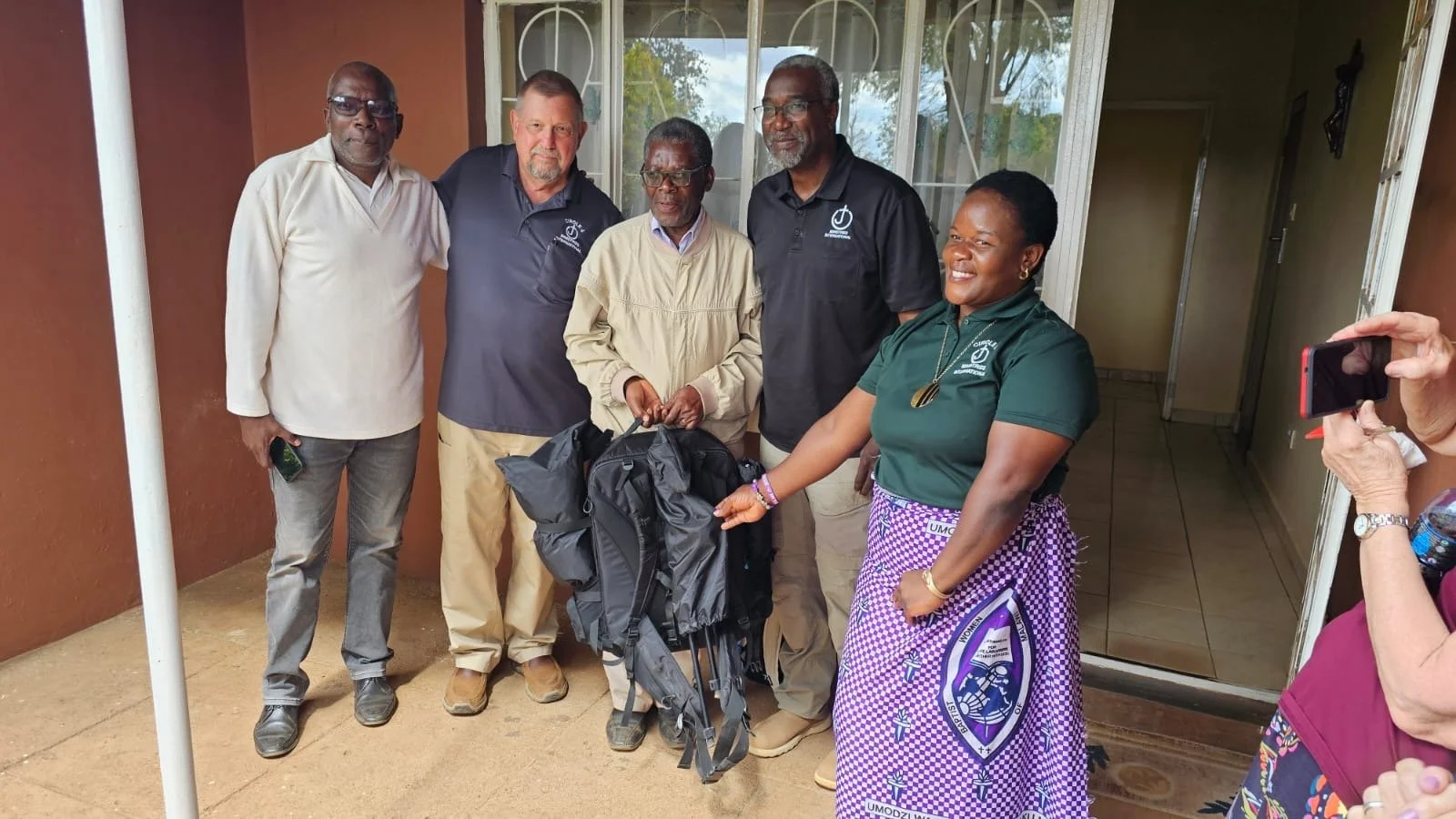 Group of six people standing outside, some smiling, including three men and one woman, with a backpack held by a man in the center. A person on the right is taking a photo.