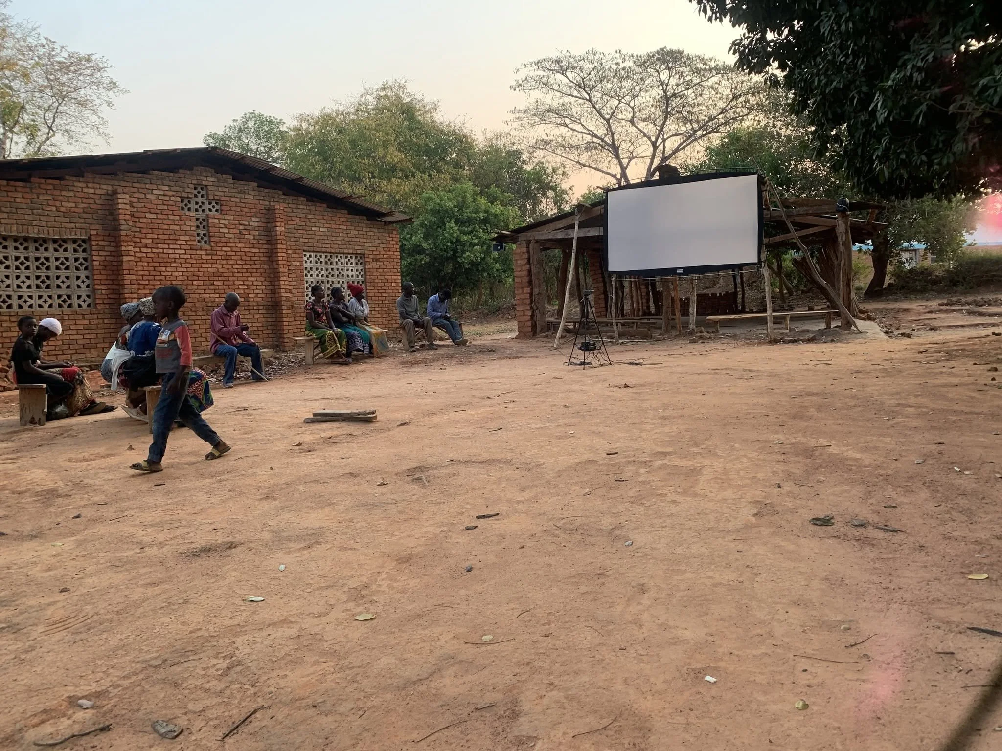 A group of people sitting on benches outside a brick building with a blank white outdoor screen set up nearby, surrounded by trees and dirt ground.