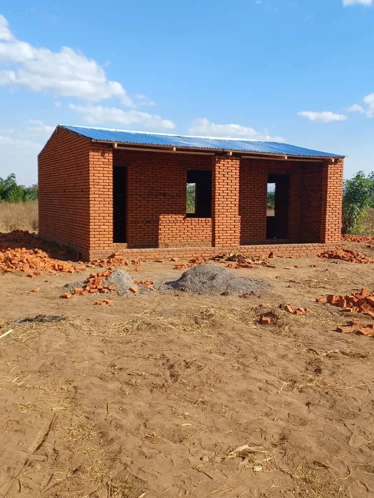A small brick house under construction with three window openings, situated on a dirt lot with construction materials and debris around, under a partly cloudy sky.