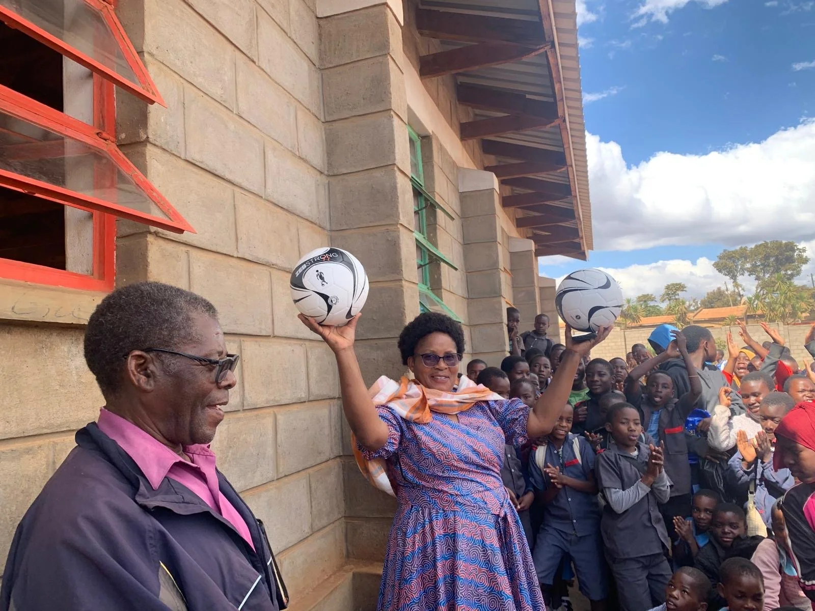 A woman holding two soccer balls in the air surrounded by a crowd of children and adults outside a building with brick walls and open windows, under a partly cloudy sky.