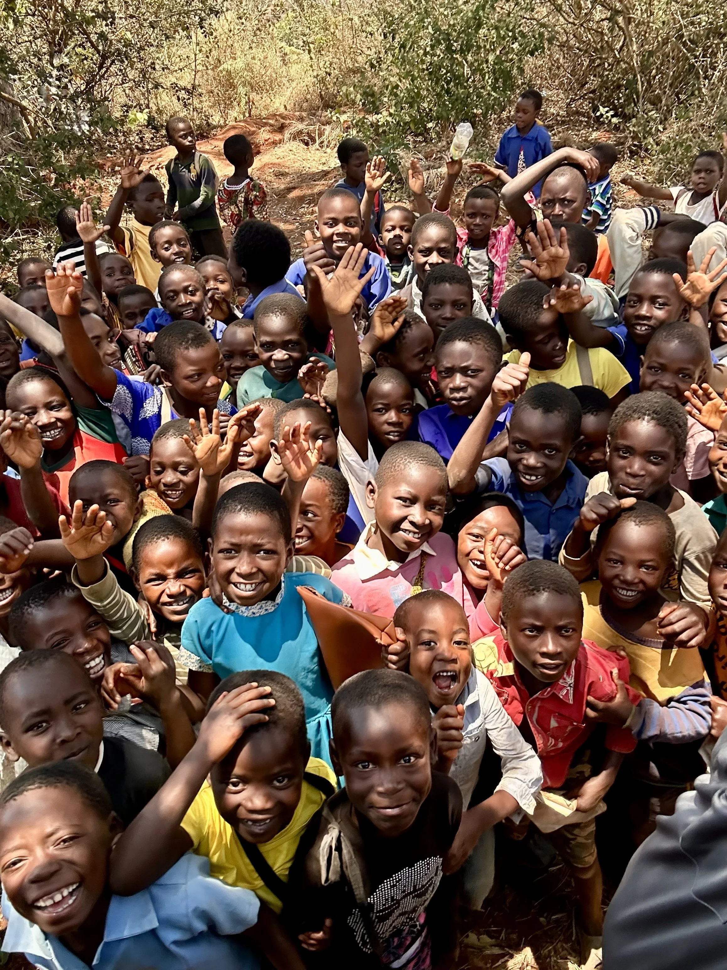 A large group of children outdoors, smiling and waving at the camera, with trees and dry ground in the background.
