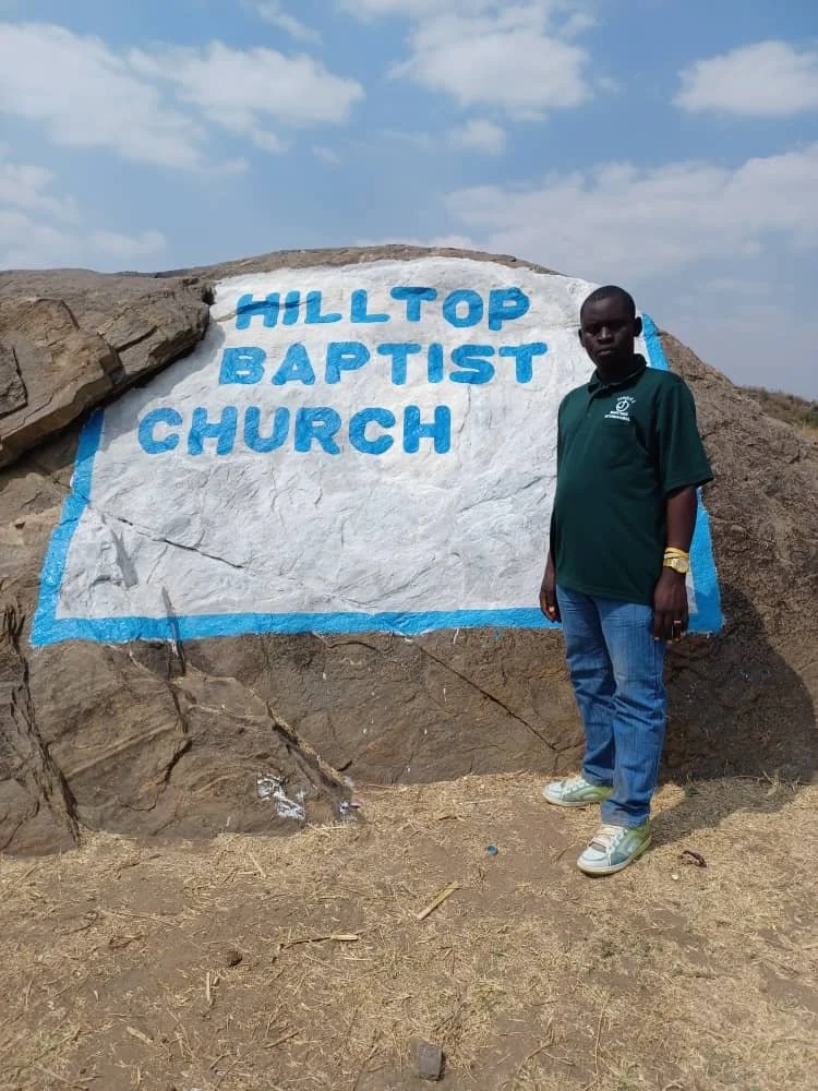 A man standing next to a painted sign on a rock that reads 'HILLTOP BAPTIST CHURCH' in blue letters.