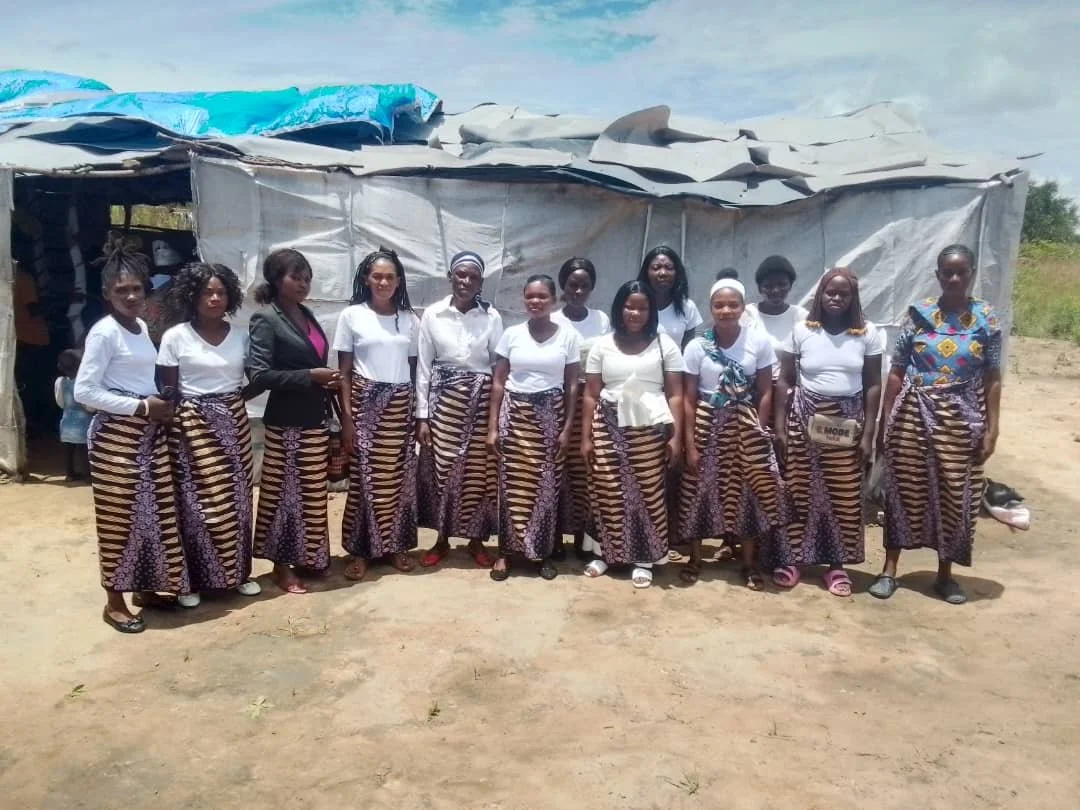 Group of women standing outdoors in front of a makeshift shelter, all wearing similar patterned skirts and white tops.