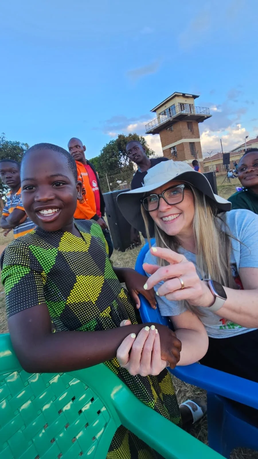 A group of children and a woman sitting outdoors on plastic chairs, smiling and interacting. The woman is wearing a wide-brimmed hat and glasses, and the children are smiling and looking at the camera. In the background, there is a watchtower structure and others standing near a fence.