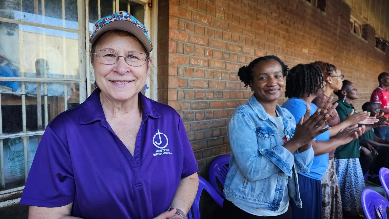 Group of women clapping at an outdoor gathering, brick wall in the background, woman in the foreground wearing a purple shirt with a logo, others wearing blue, green, and patterned clothing.