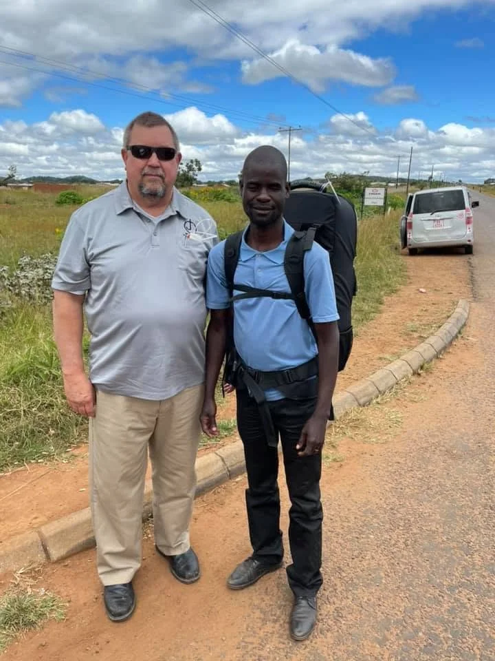 Two men standing on roadside, one in a gray polo and khakis, the other in a blue polo with a large backpack, under a partly cloudy sky.
