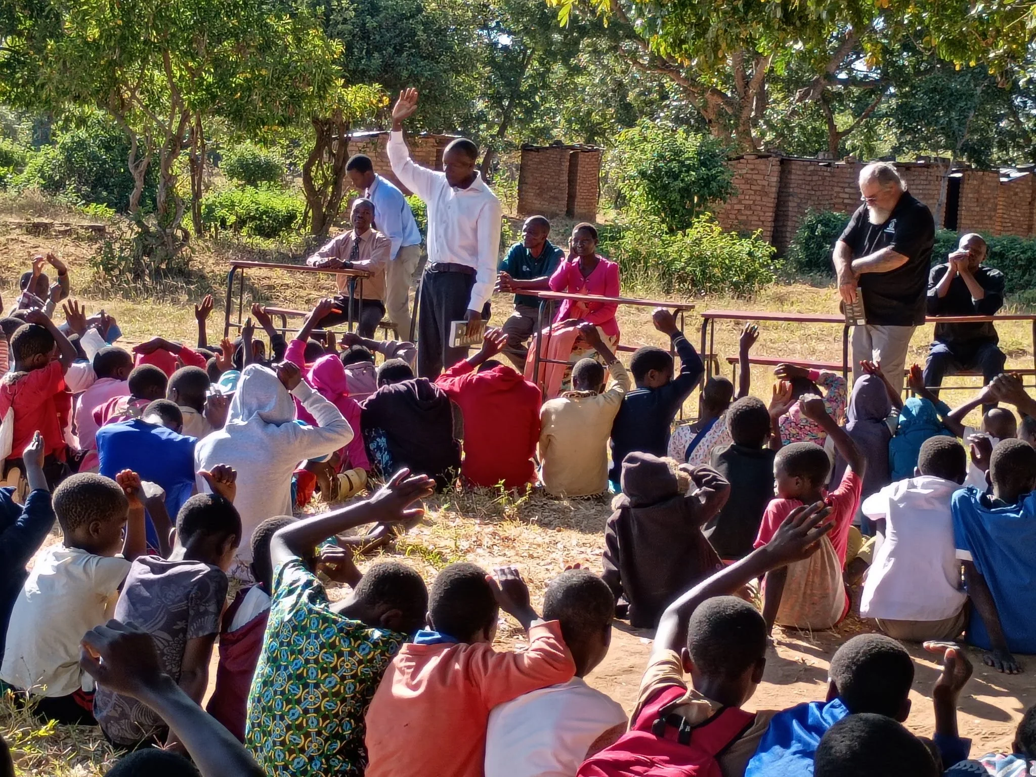 A group of children sitting on the ground in front of a stage in an outdoor setting with trees and brick walls, watching and raising their hands. On the stage, there are five adults, one of whom is standing and waving, others seated or standing, with one holding a book. The scene appears to be a community gathering or event.