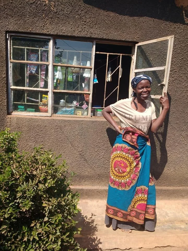 A smiling woman standing in front of a brown textured wall with a window, holding a window frame, and wearing a patterned skirt, headscarf, and t-shirt.
