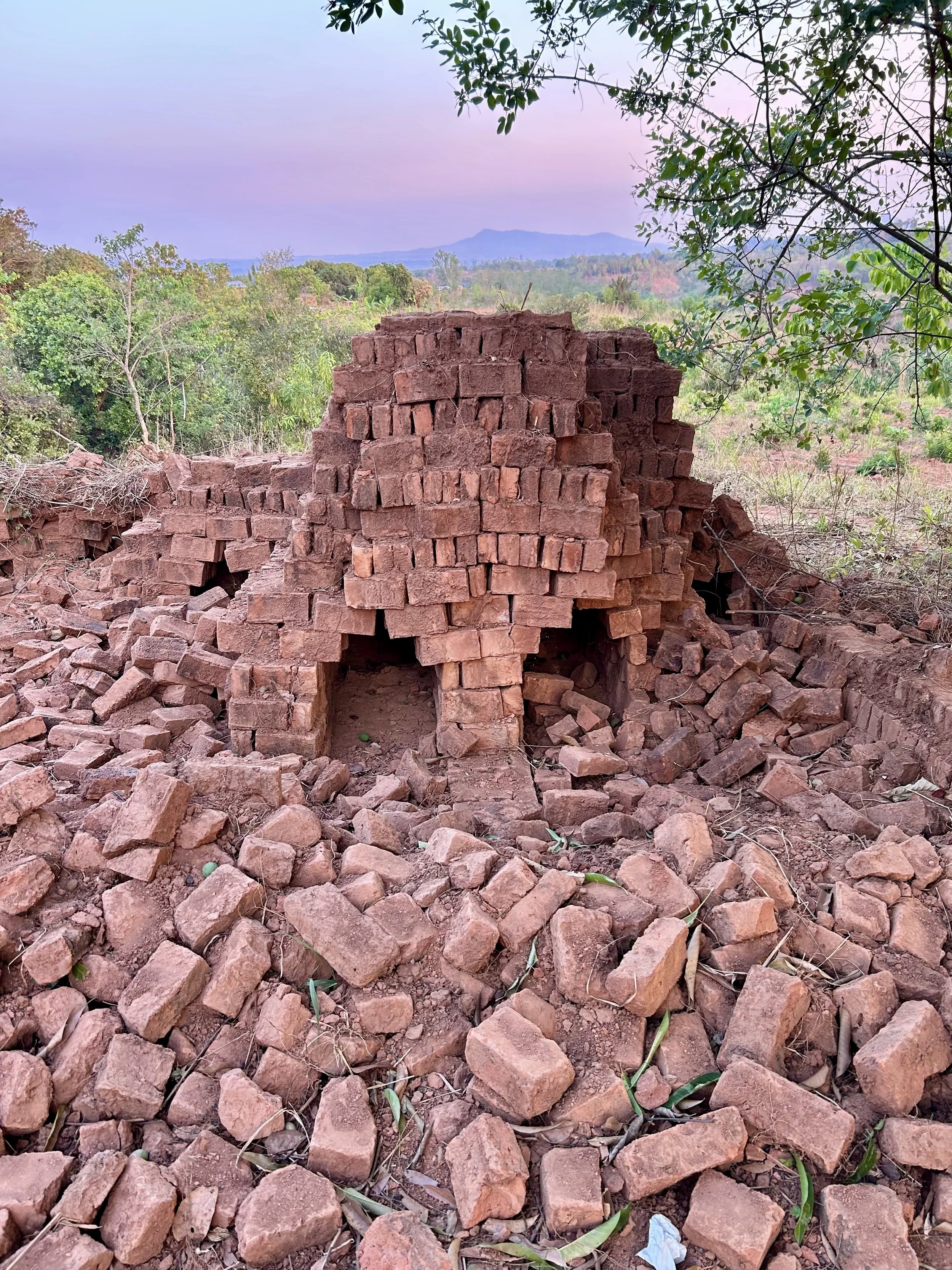 A partially collapsed brick structure in a rural area with scattered bricks around. Tree branches and green foliage are visible in the foreground, with a scenic view of distant mountains and a pastel sky in the background.