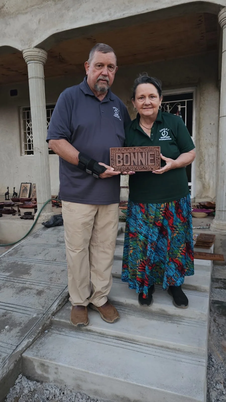 A man and woman holding a wooden sign that says 'BONNE' in front of a house with columns. The woman is wearing a green shirt with a logo and a colorful skirt, and the man is wearing a navy shirt and beige pants.