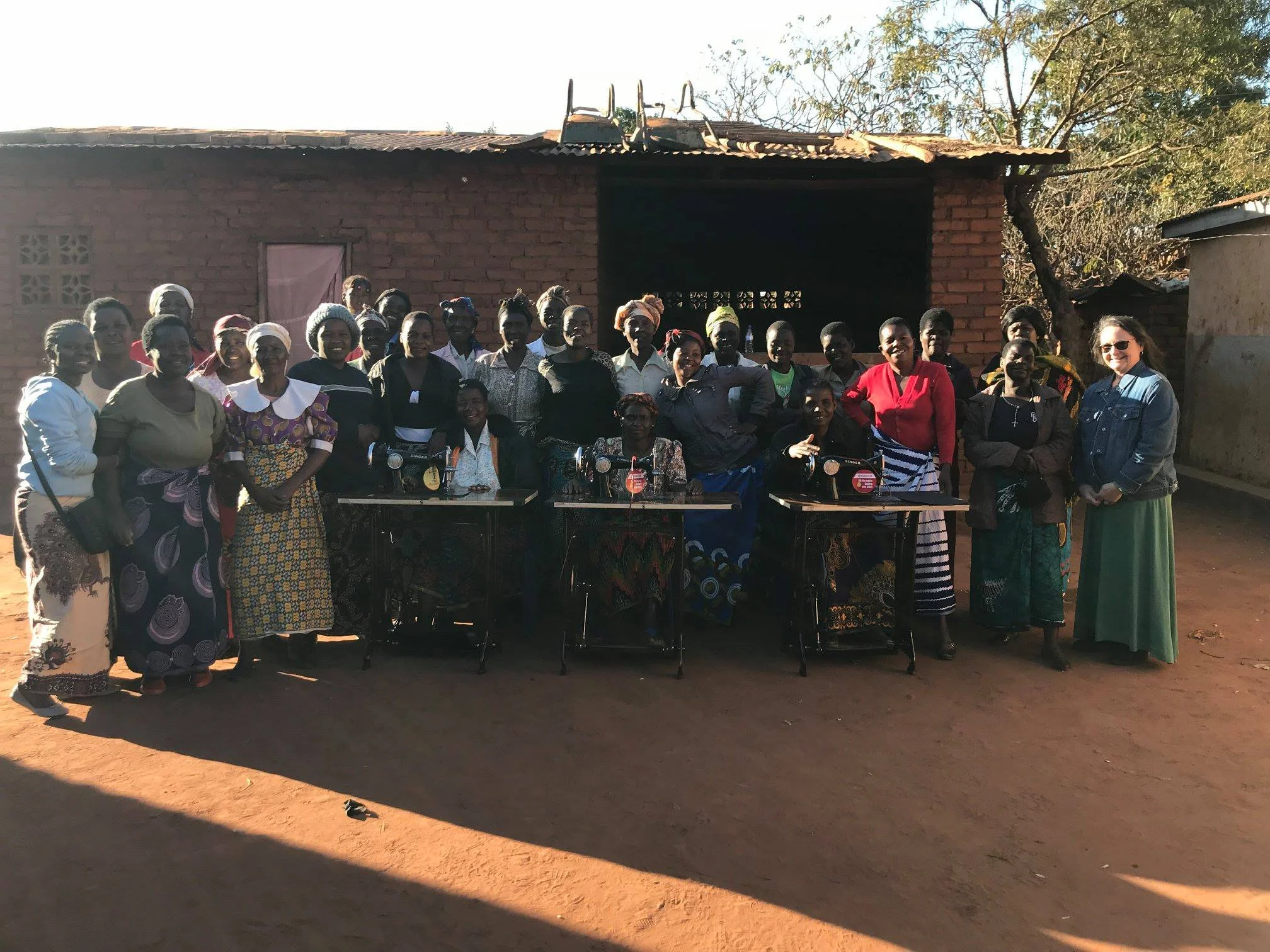 Group of women and two women on the right with sunglasses standing in front of a brick building, some smiling, some with neutral expressions, during daylight in a rural setting.