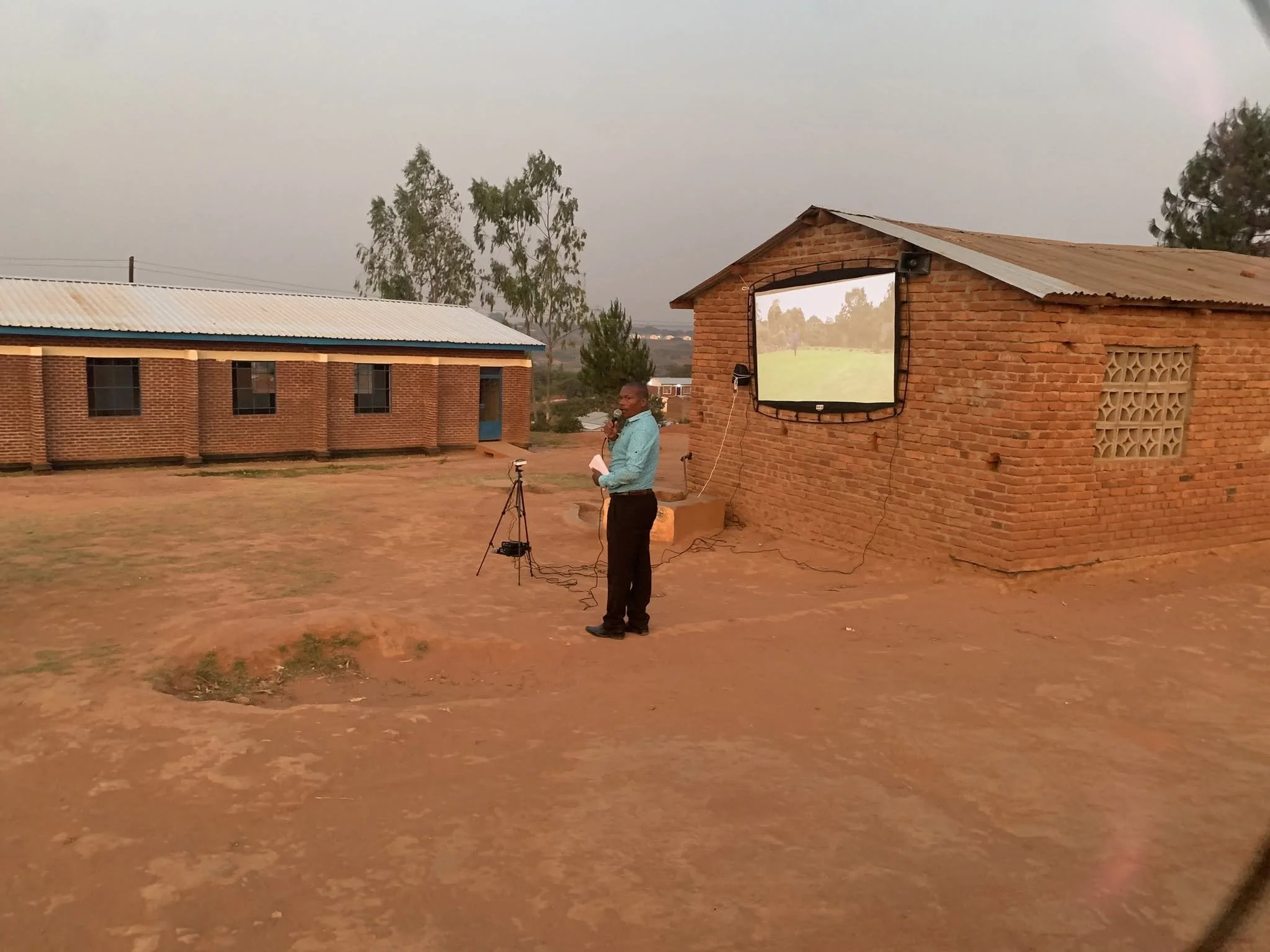 A man stands outdoors speaking into a microphone, with a projection screen displaying a landscape scene behind him. The area has brick buildings and a few trees, and the sky is overcast.