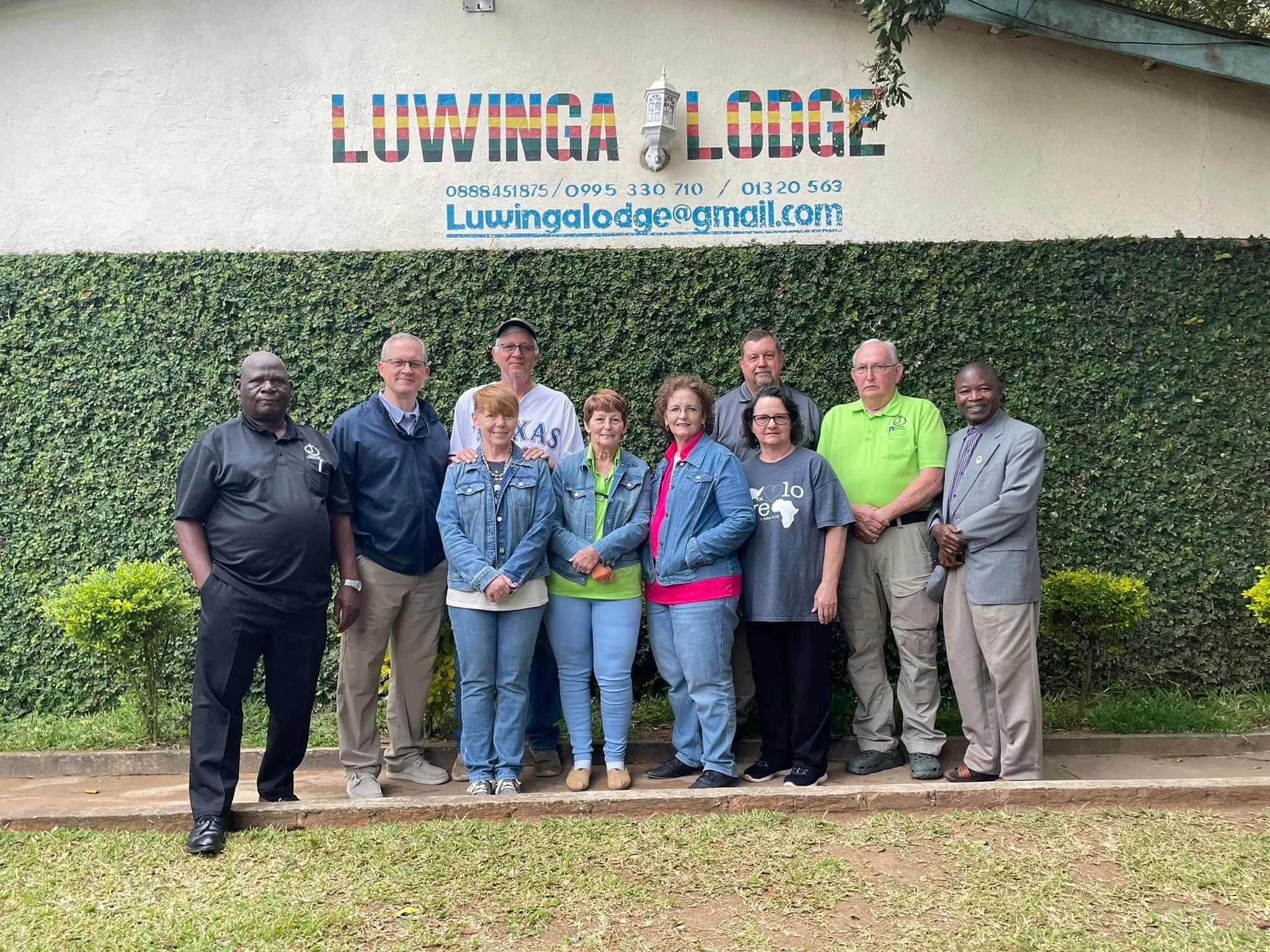 A group of eleven people standing outdoors in front of a wall with a colorful sign reading 'LIVINGA LODGE' and a contact email. The group is diverse in age and appearance, with some wearing glasses, casual clothing, and smiling at the camera.
