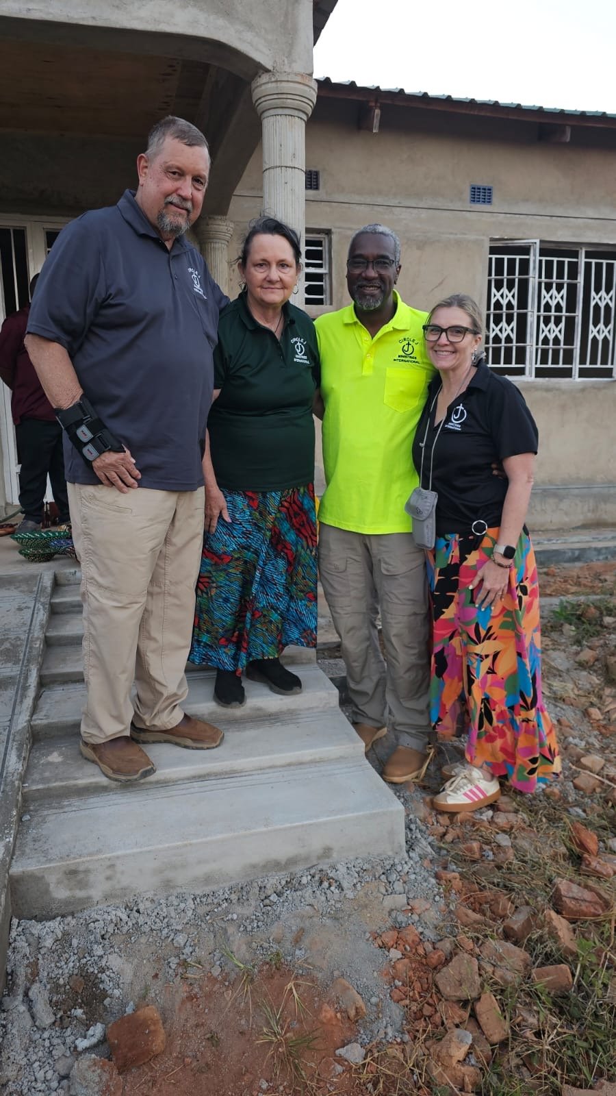 Four people standing on front steps of a building, smiling for the photo. Two men and two women, dressed casually with some wearing colorful skirts and t-shirts with logos.
