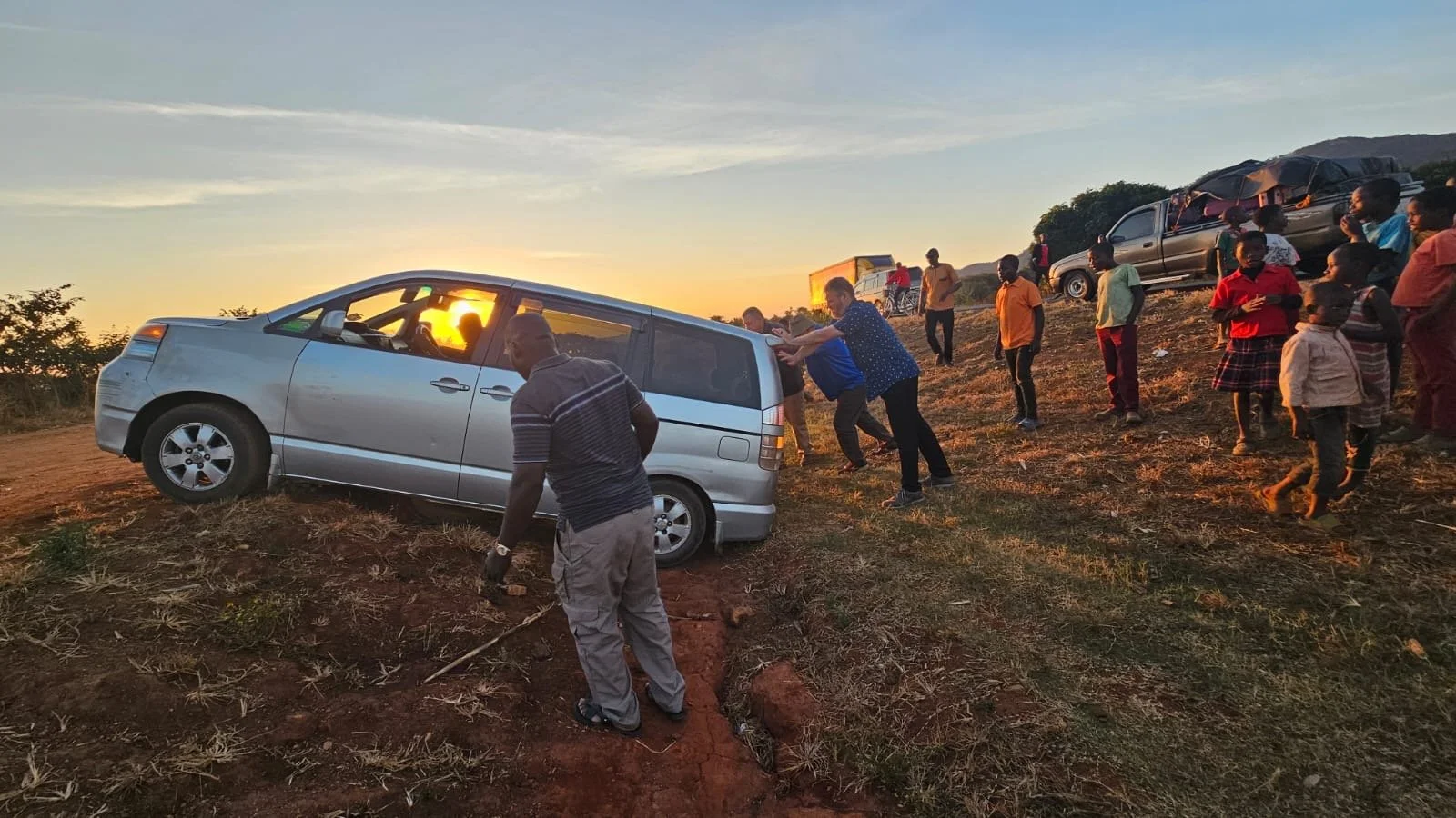 People helping a silver minivan stuck on a dirt hill during sunset, with several other vehicles and children in the background.