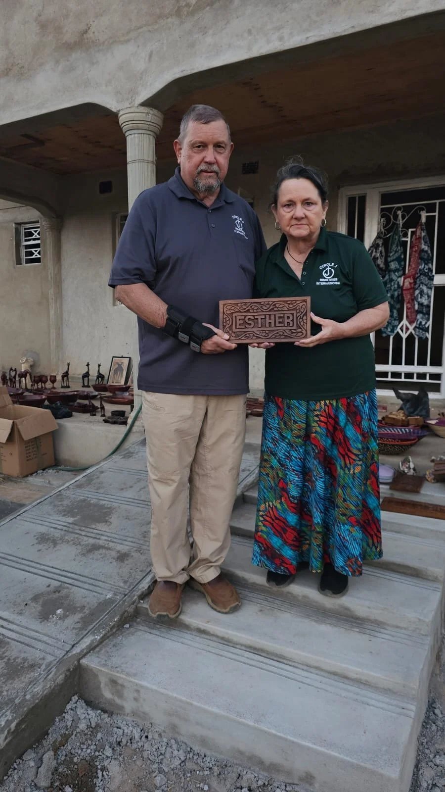A man and woman standing on a porch, holding a wooden plaque with the name 'Esther', in front of a house with various decorations and items displayed on the ground.