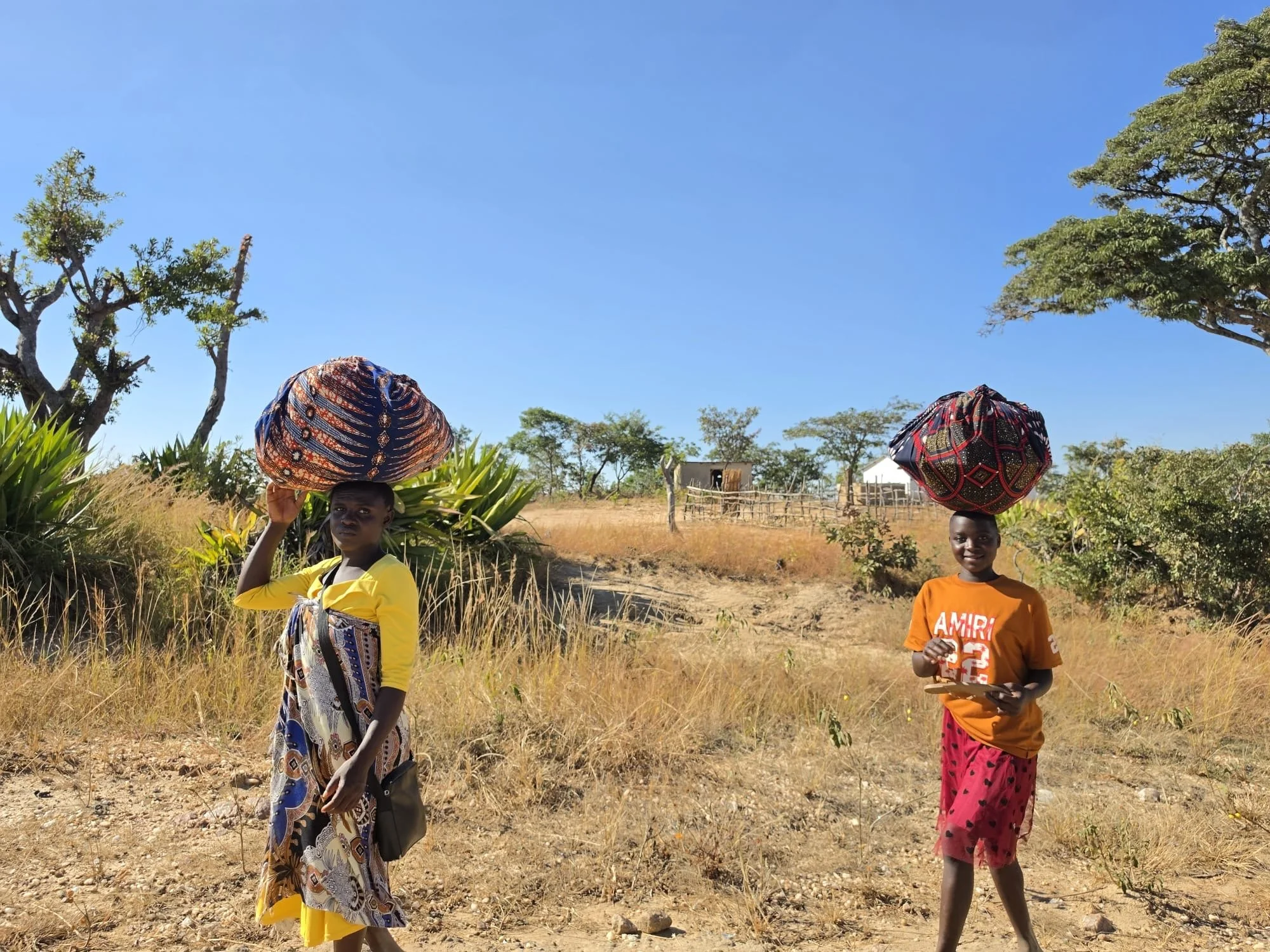 Three children standing outdoors on dry grassy ground, each balancing a large, colorful, patterned basket on their heads. Two girls are smiling at the camera, with trees and a small fence in the background under a clear blue sky.