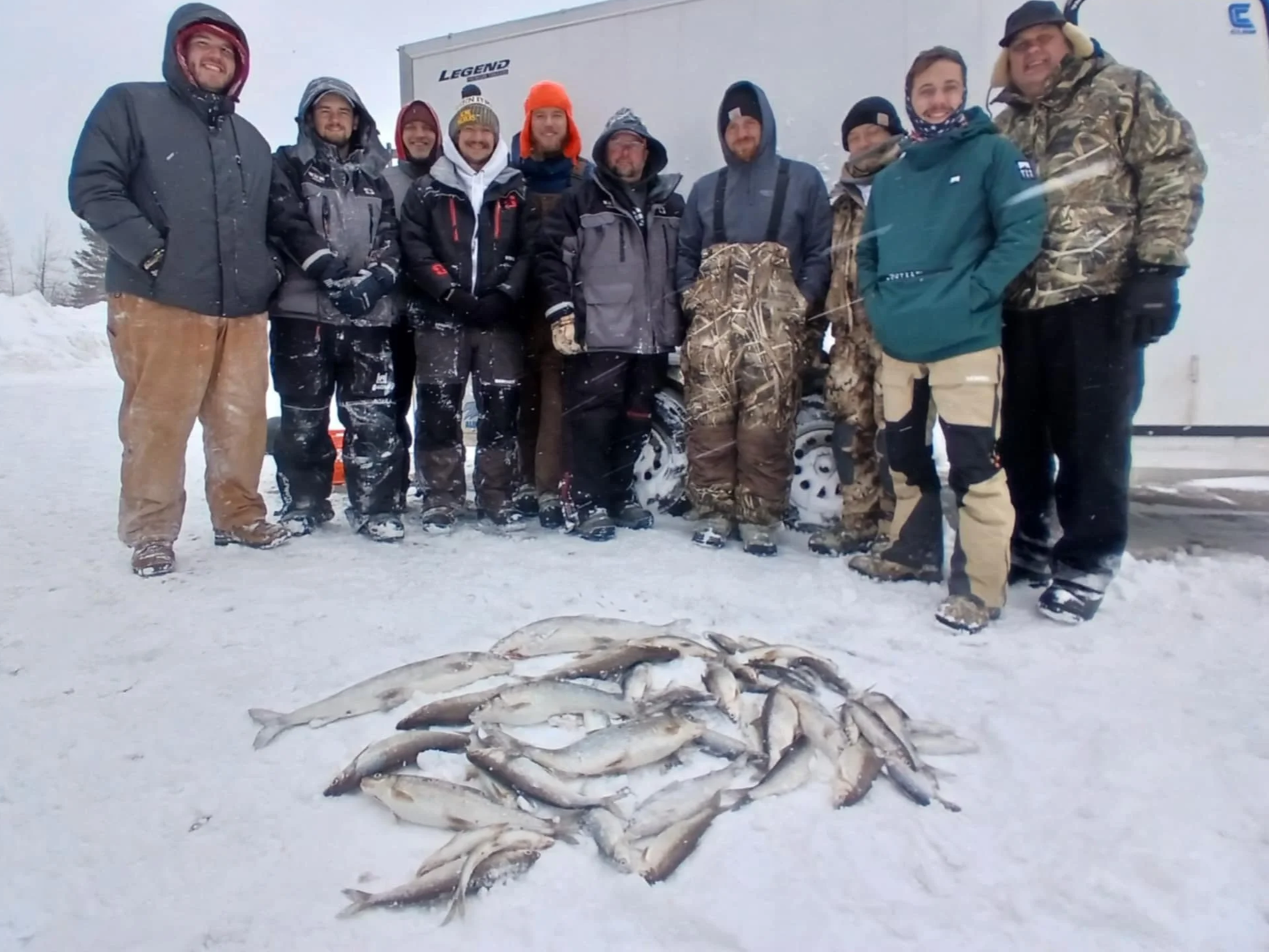 A family ice fishing on Lake Superior, holding various fish, with more fish on the snowy ground, and a blue ice shelter behind them.