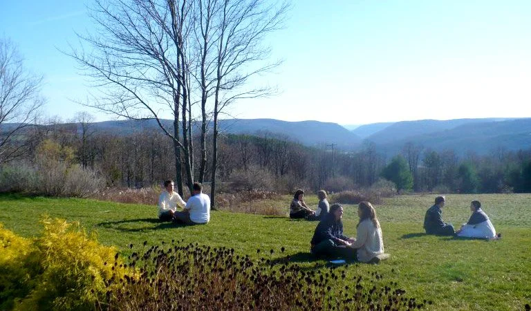 People sitting in pairs on a grassy hillside with mountains and leafless trees in the background.