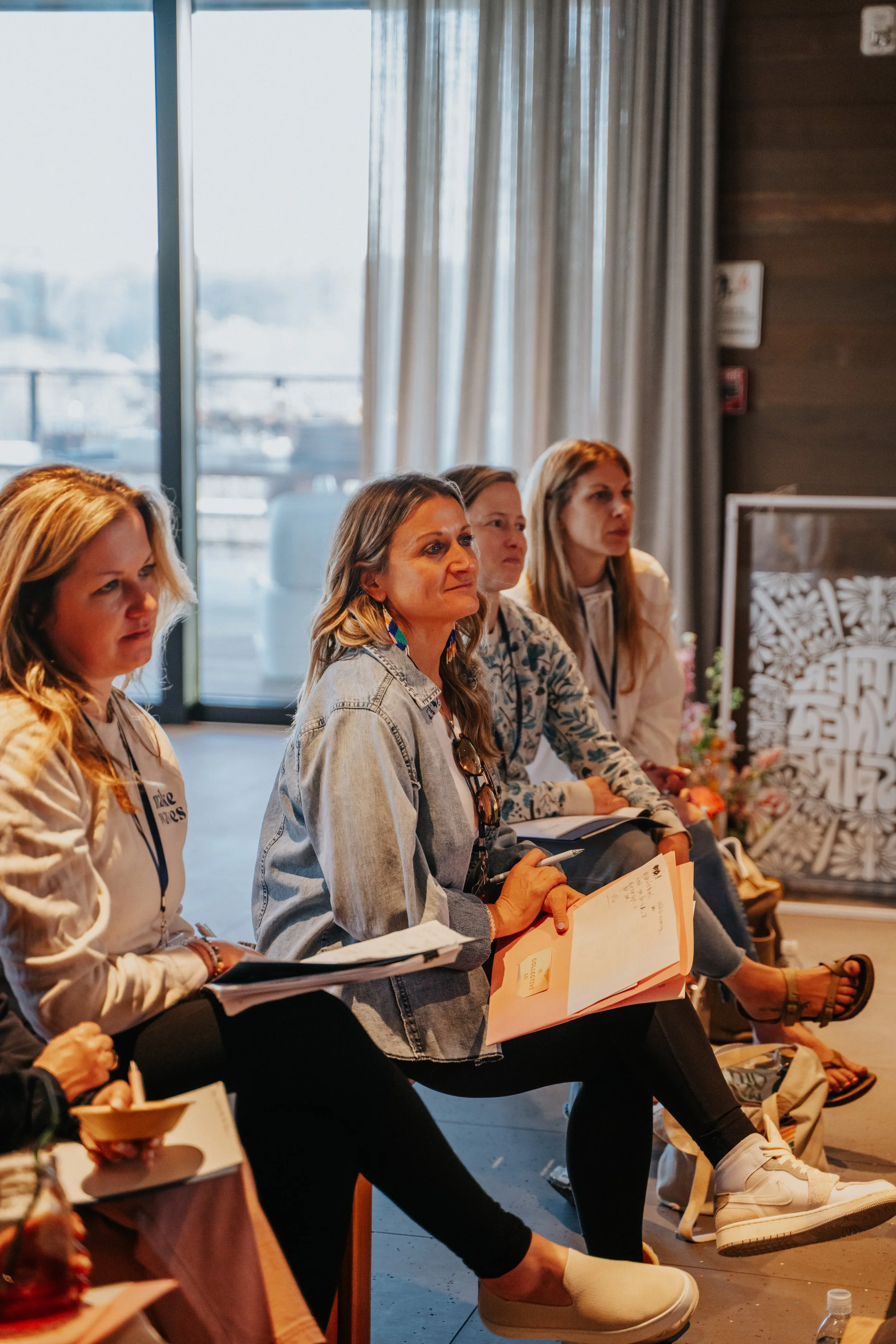 Four women sitting in a row appears to be at an indoor event, holding notebooks and pens, with a large window and curtains in the background.
