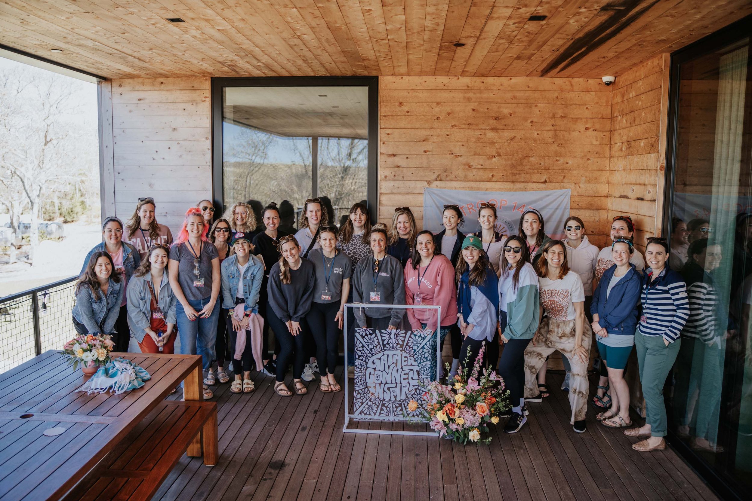 Group of women gathered on a wooden balcony for a photo, with a banner in the background and a bouquet of flowers on a table in the foreground.