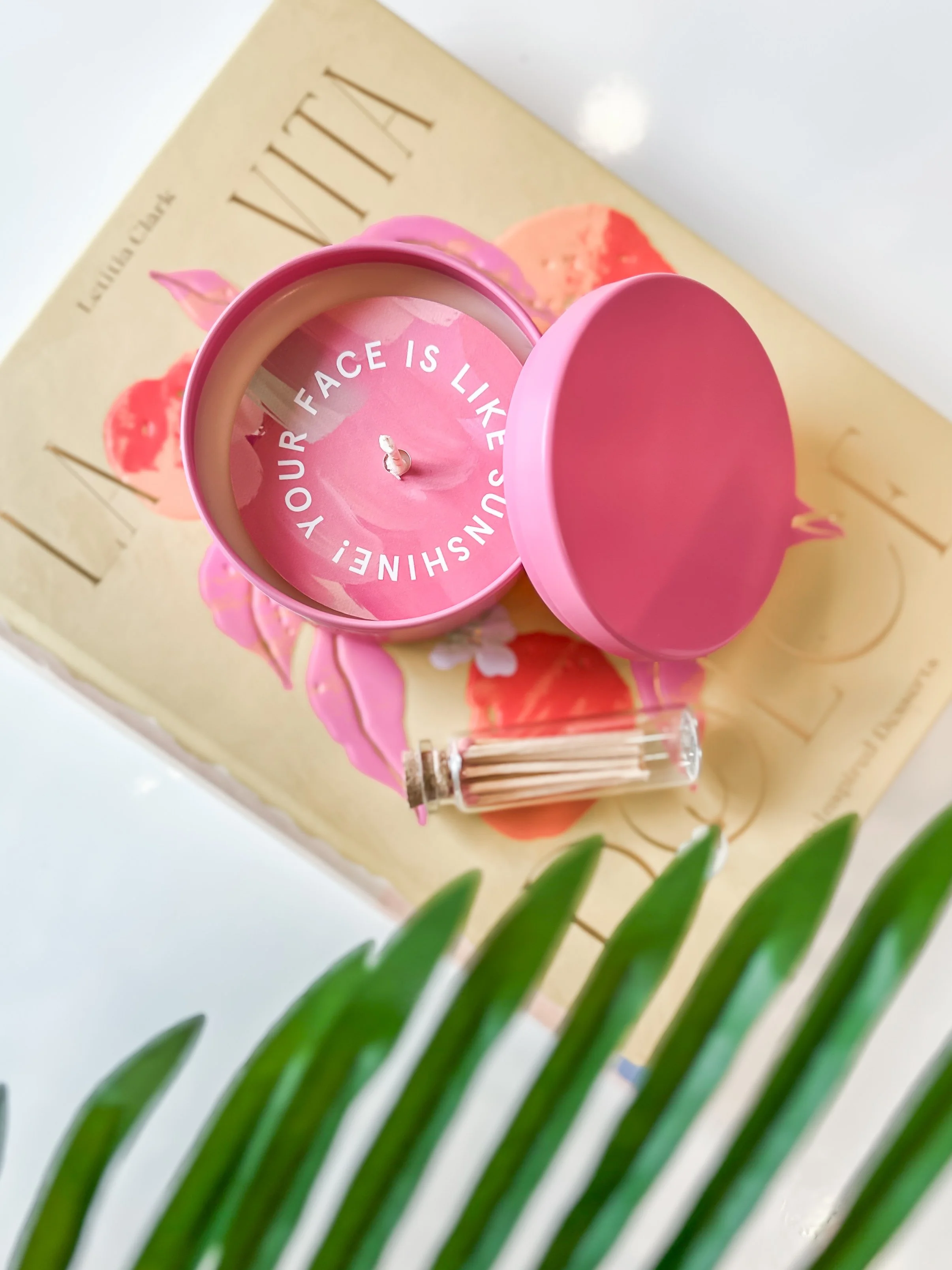 Close-up of a pink candle with the words 'Your face is like sunshine' inside the lid, with a floral background, a book titled 'VITAMIN', and a plant in the foreground.