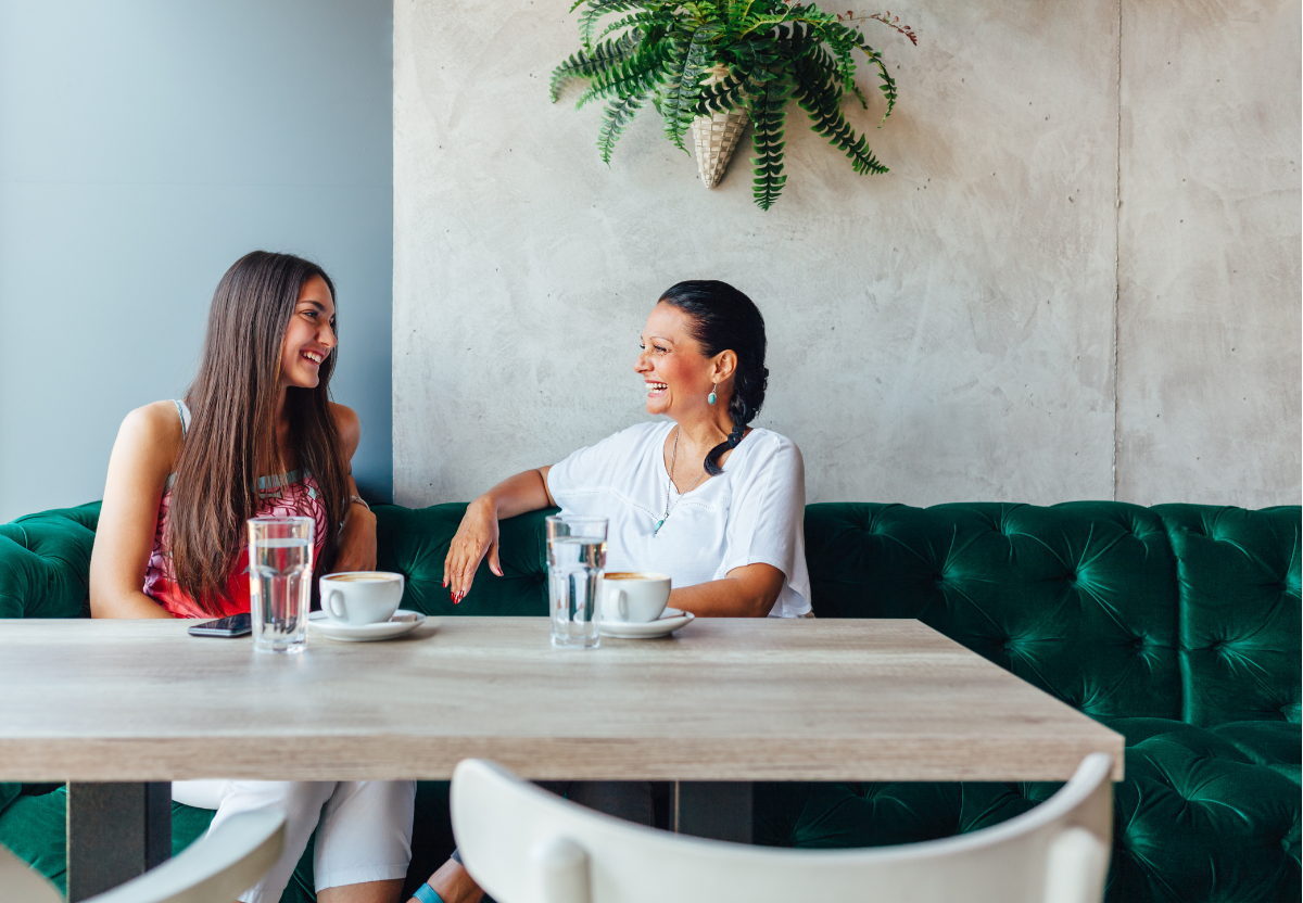 Two women sitting at a table in a cafe, laughing and talking. The table has two cups of coffee, two glasses of water, and a smartphone. A potted fern hangs on the wall behind them.