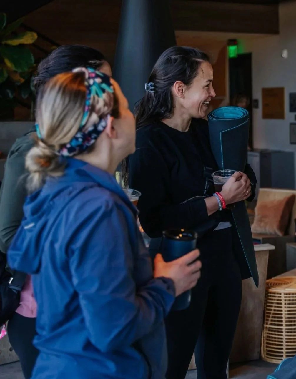 A group of people smiling and talking indoors, one woman holding a yoga mat and a glass of water.