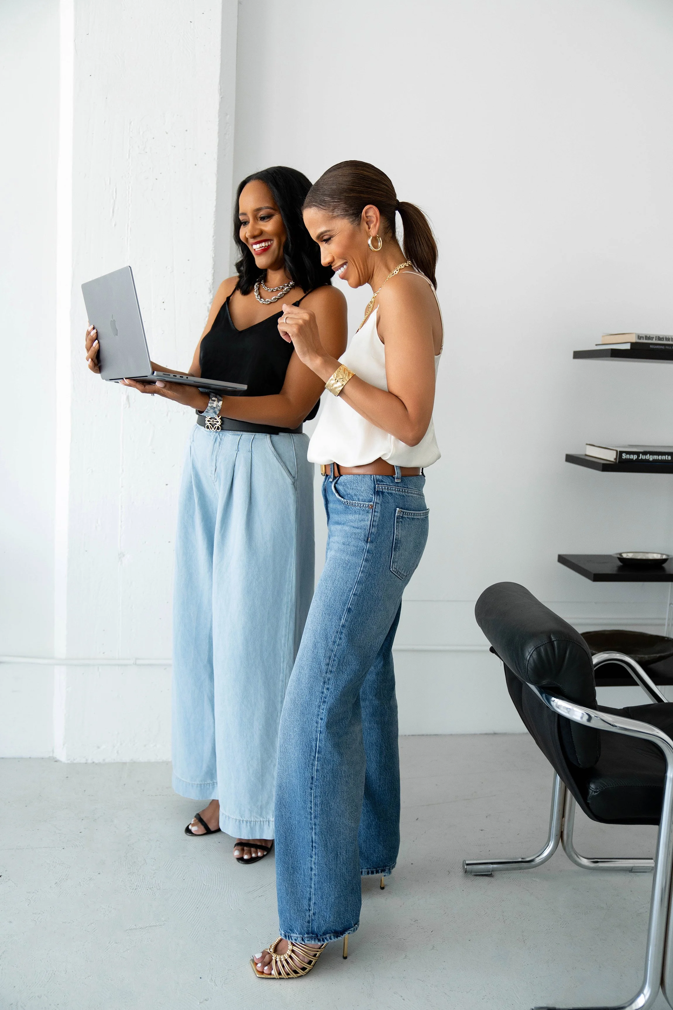 Two women standing and looking at a laptop computer in a minimalist room with white walls and a black chair.
