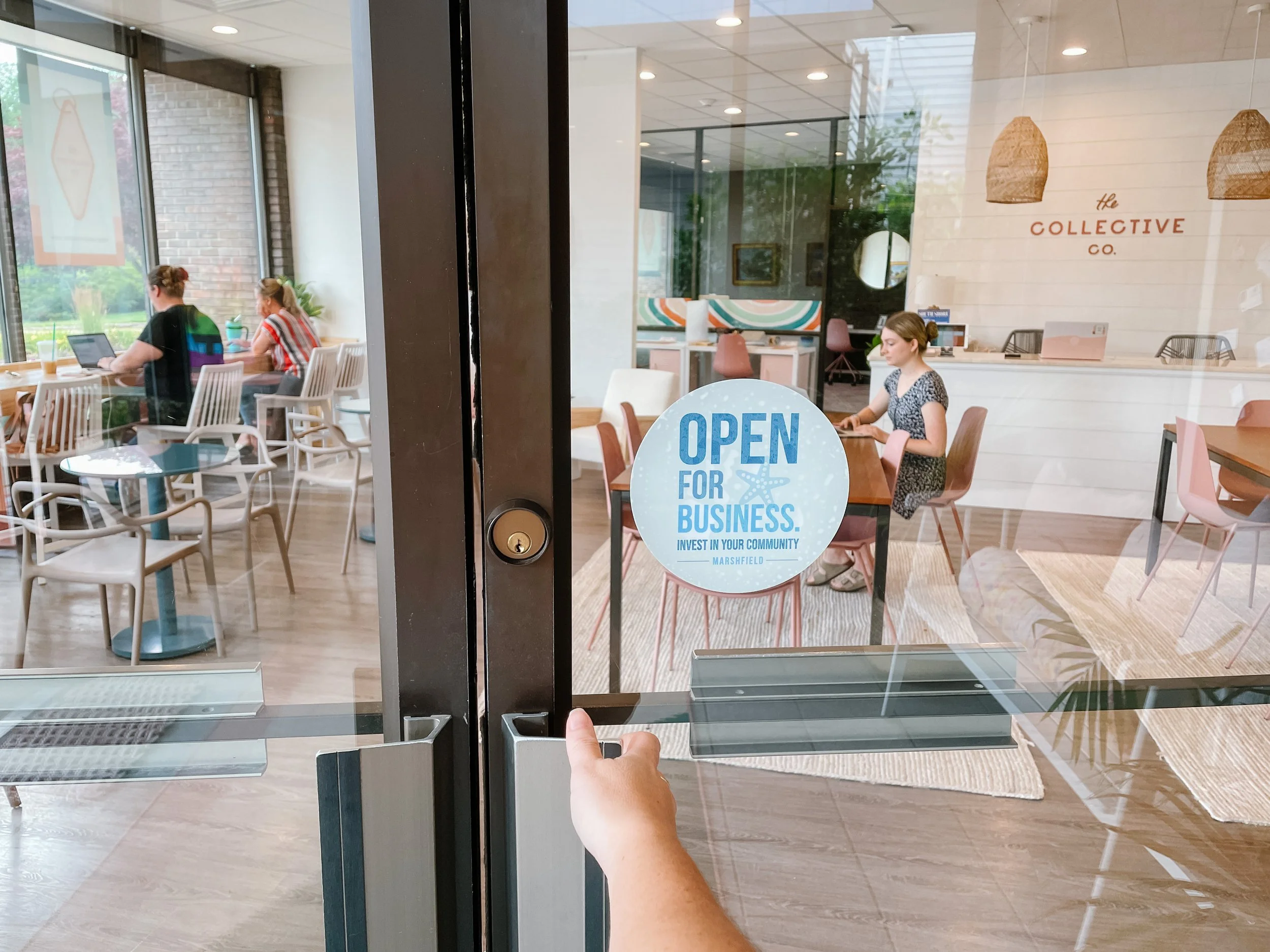 A person pressing a glass door release button inside a cafe, with an 'Open for Business' sign on the door. The cafe interior has tables, chairs, and customers working on laptops.