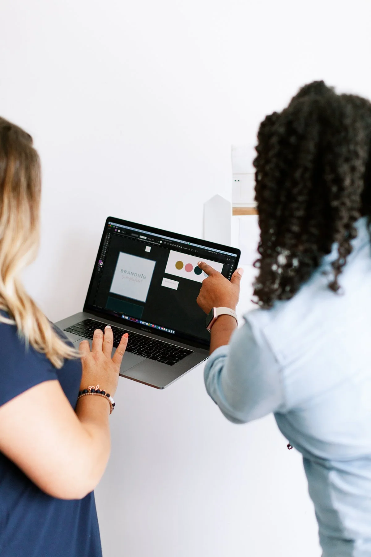 Two women working on a laptop with a design project on the screen in an office with plain white walls.